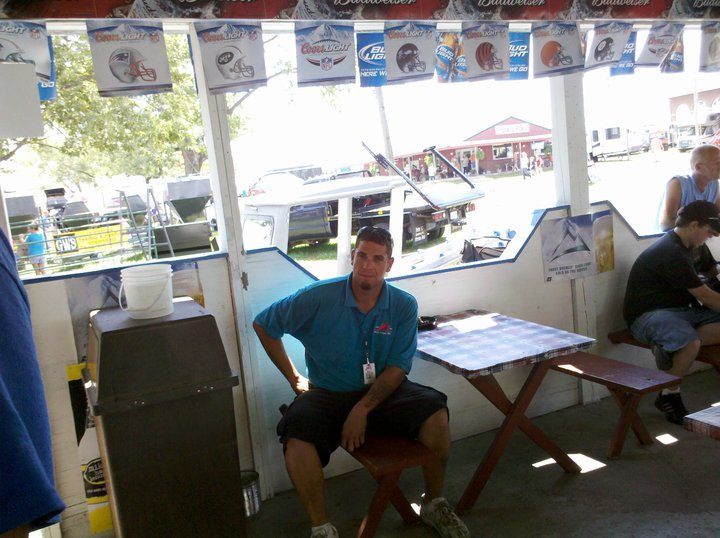 A man in a blue shirt sits on a bench inside a food stand, with NFL team banners above and picnic tables around. The background shows a sunny outdoor event.