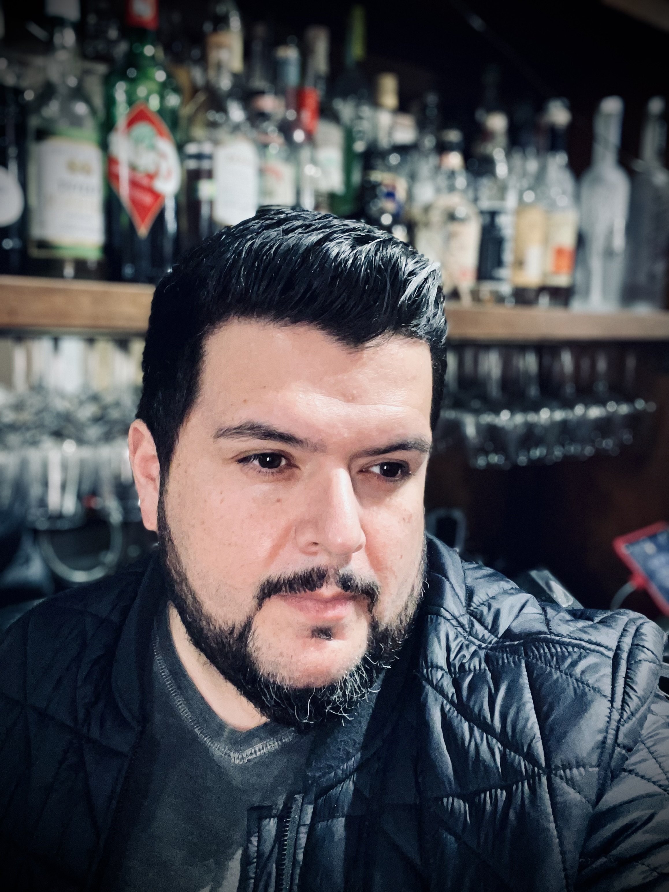 A person with dark hair and a beard is looking to the side in a dimly lit bar, with shelves of various liquor bottles and rows of hanging glasses in the background.