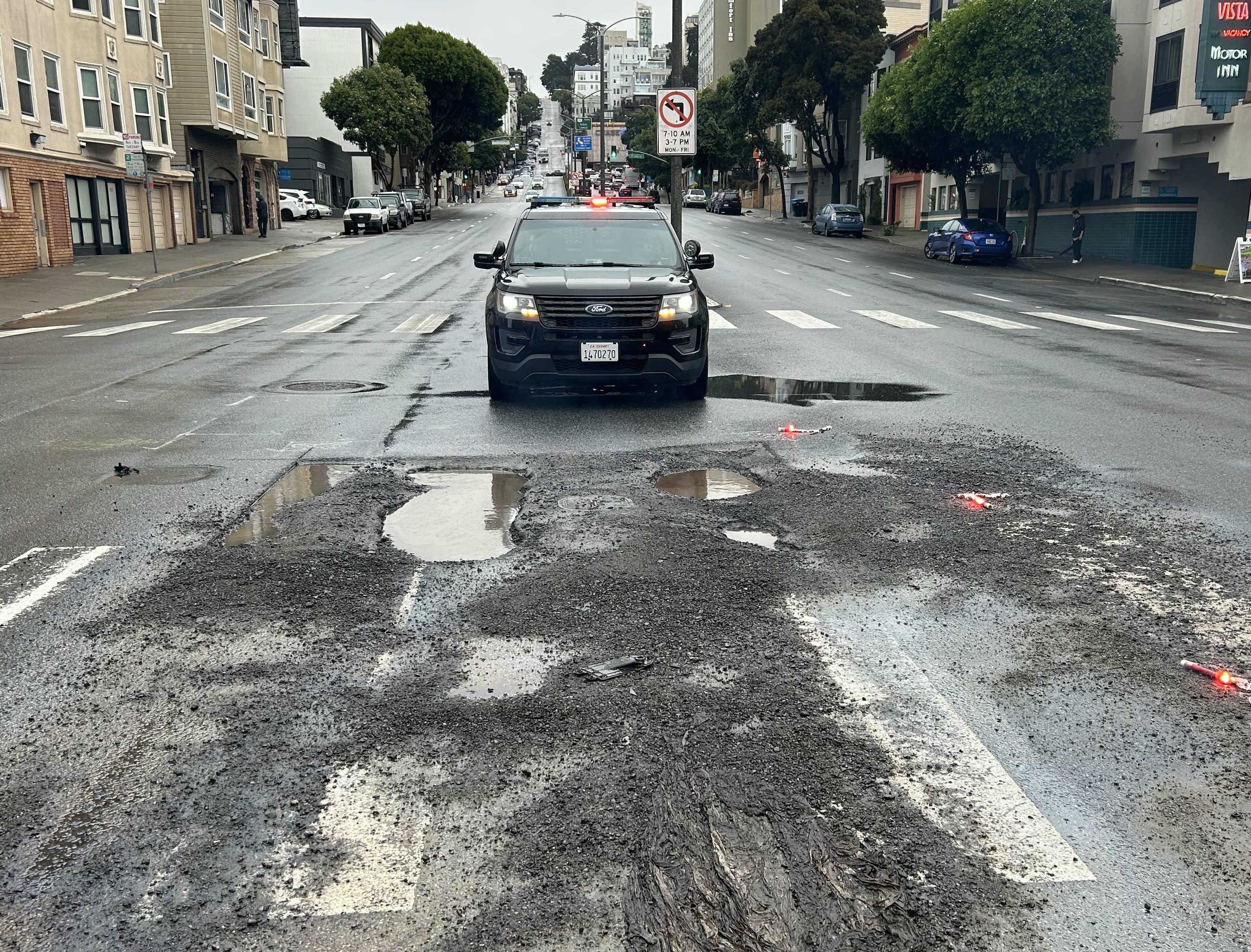 A black vehicle approaches a messy intersection, with wet road and debris scattered. Traffic lights are red, and trees line the street in the background.