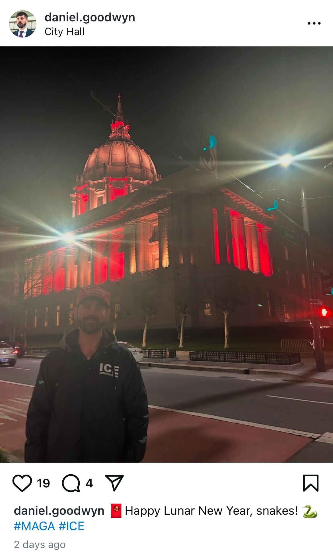 A man stands on a street at night in front of a city hall building, which is illuminated with red lights. He wears a dark jacket with &quot;ICE&quot; printed on it.