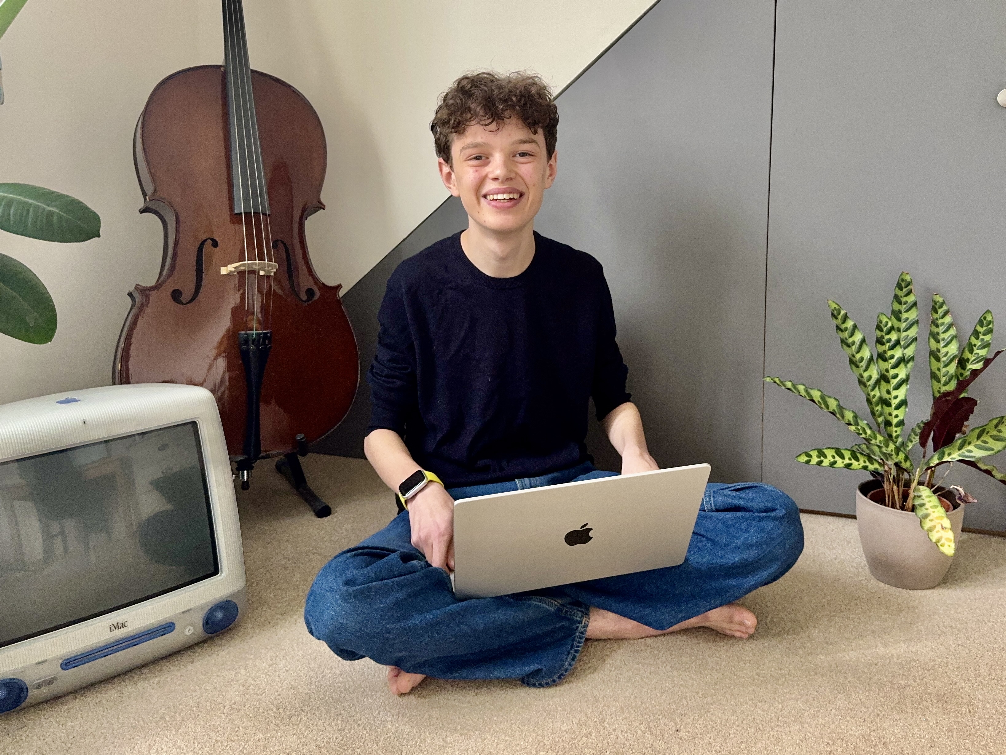 A person sits cross-legged on the floor, smiling with a laptop. Nearby are a cello, a vintage iMac, and a potted plant, creating a cozy, mixed setting.