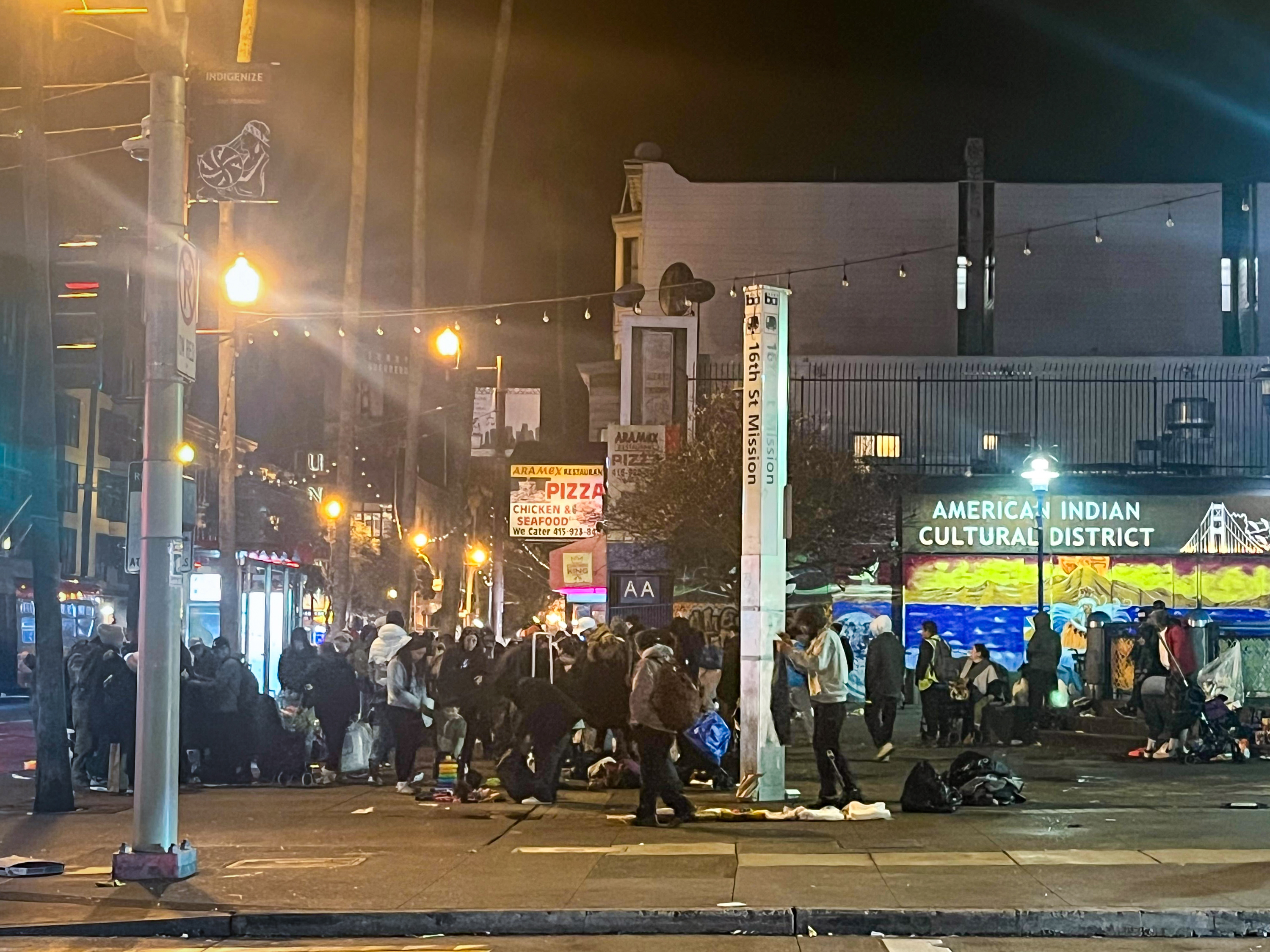 A nighttime street scene with a group of people gathered on a sidewalk. Signs and streetlights illuminate the area, featuring a mural for the &quot;American Indian Cultural District.&quot;