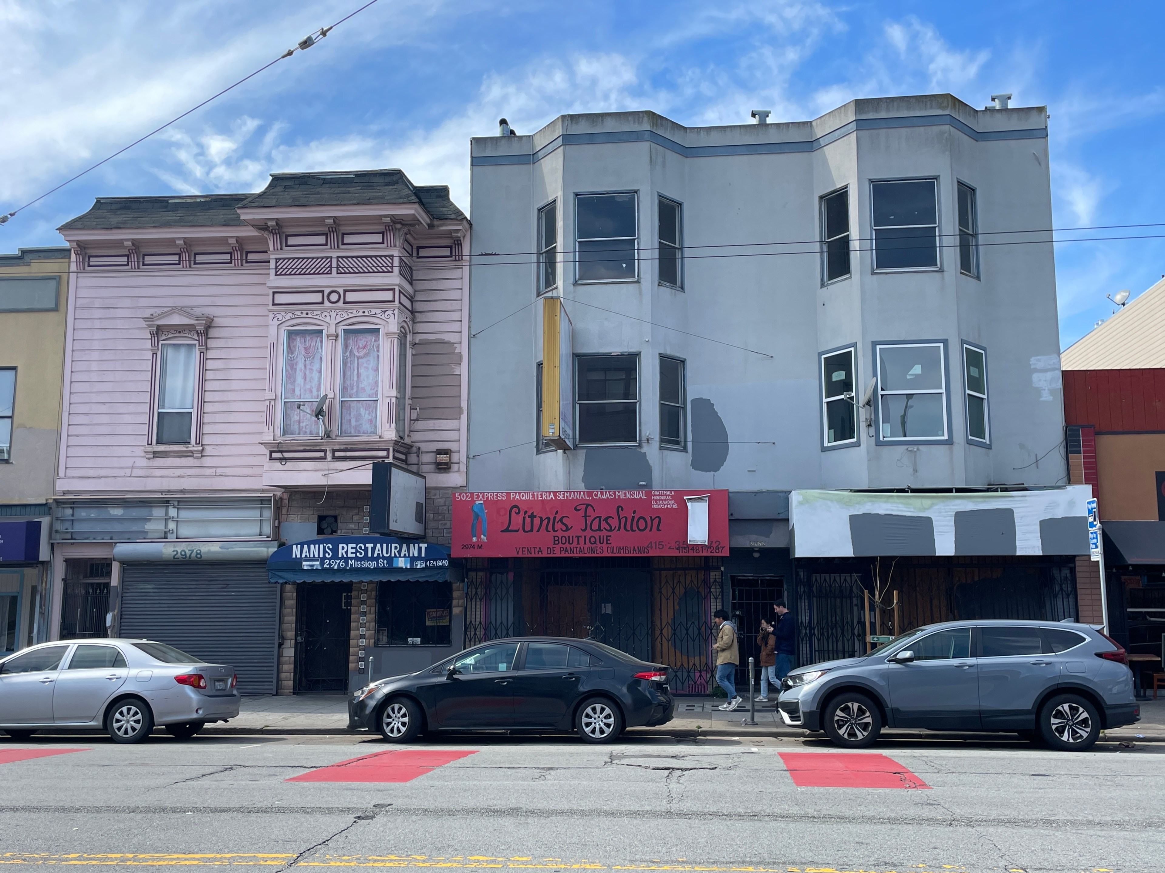 The image shows a street with three cars parked in front of buildings. There's a pink Victorian-style building beside a gray, modern one, with shops below.