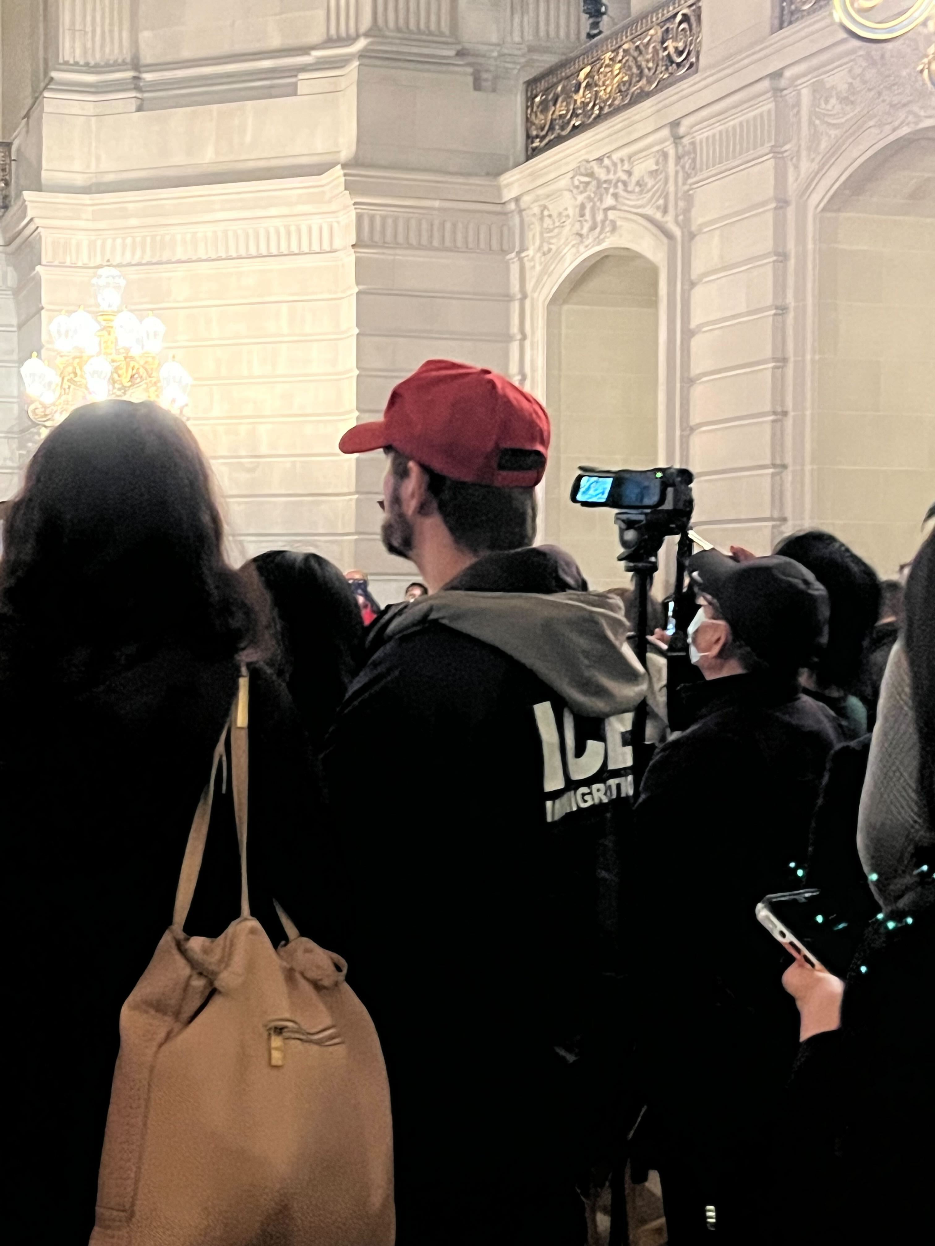 A group of people, including a person in a red cap with an &quot;ICE&quot; jacket, stand in an ornate room with a tripod camera and chandelier.