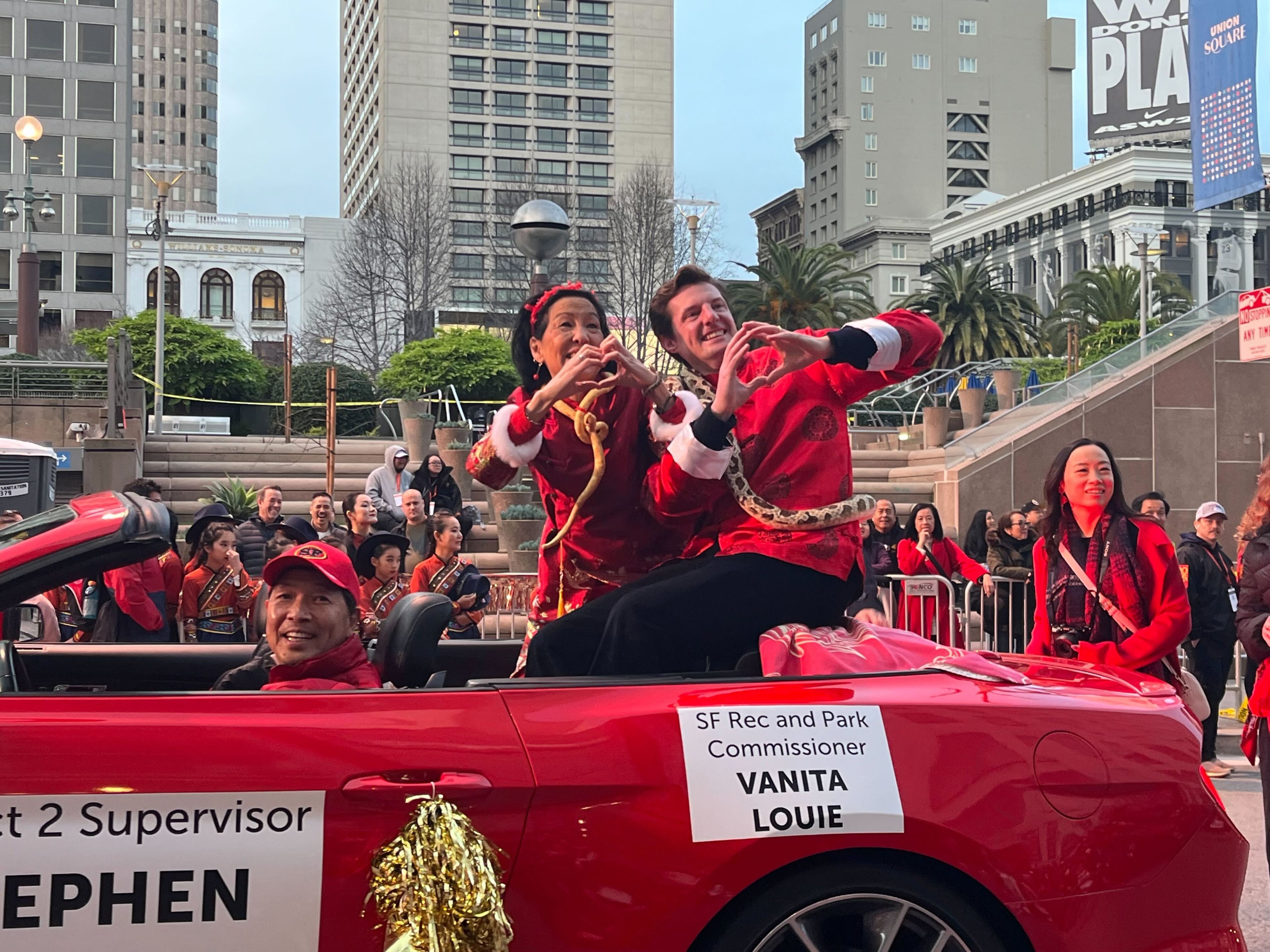 Two people in Santa outfits are smiling and making a heart shape with their hands while sitting in a red convertible. A crowd is gathered around them.