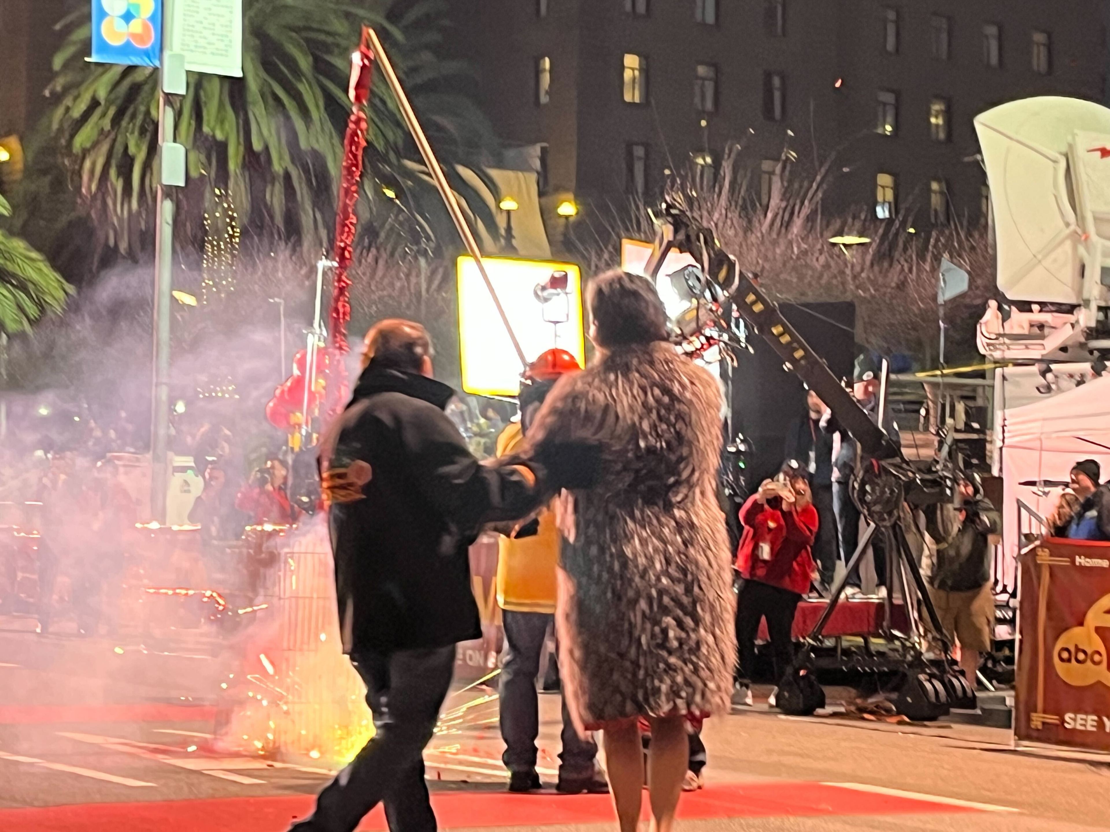 Two people stand on a red carpet surrounded by smoke and bright lights. A camera crew films near a building, and there are festive decorations and a palm tree.