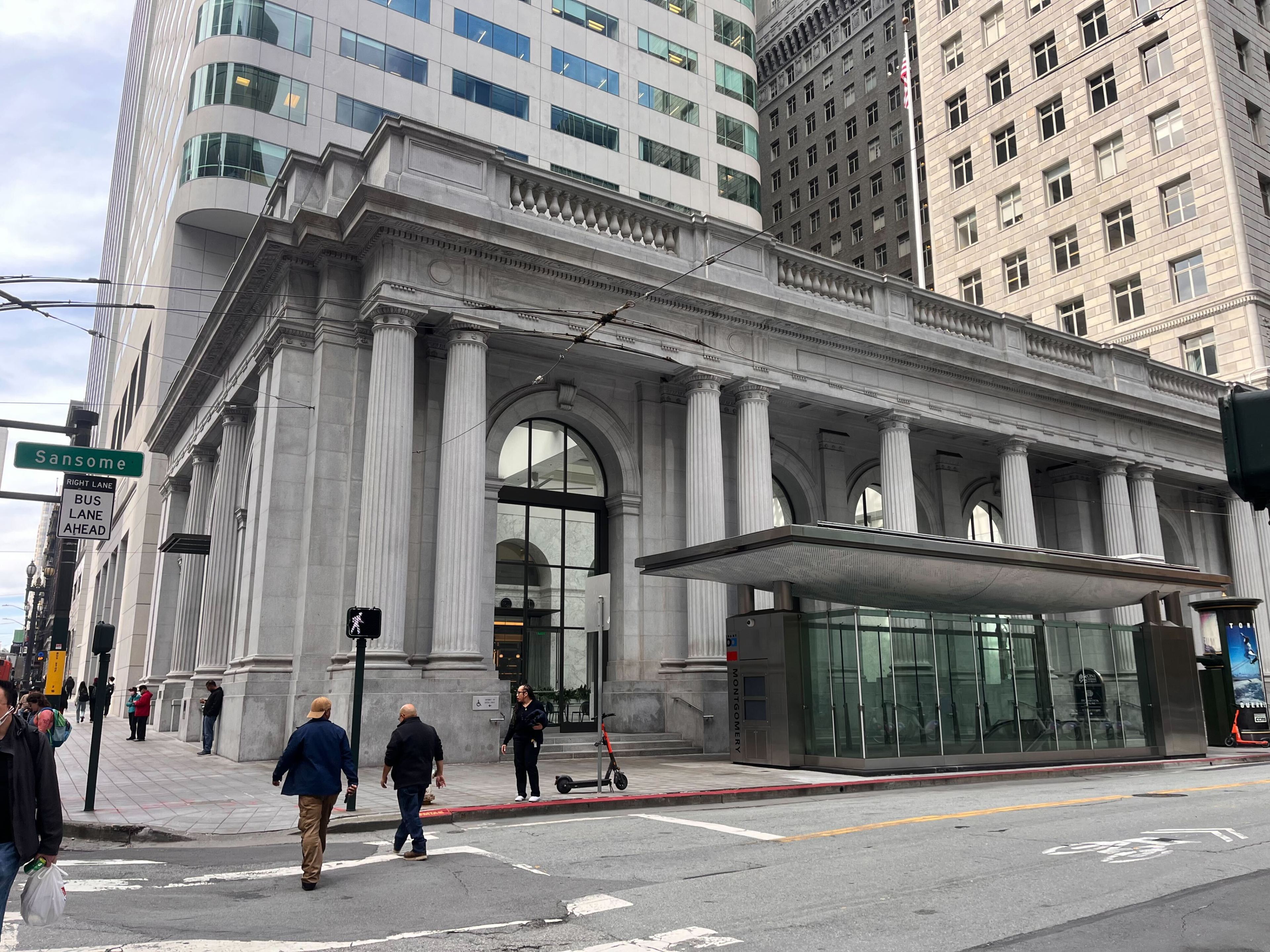 A neoclassical building with tall columns stands on a street corner, surrounded by modern skyscrapers. People walk along the sidewalk next to a glass entrance.