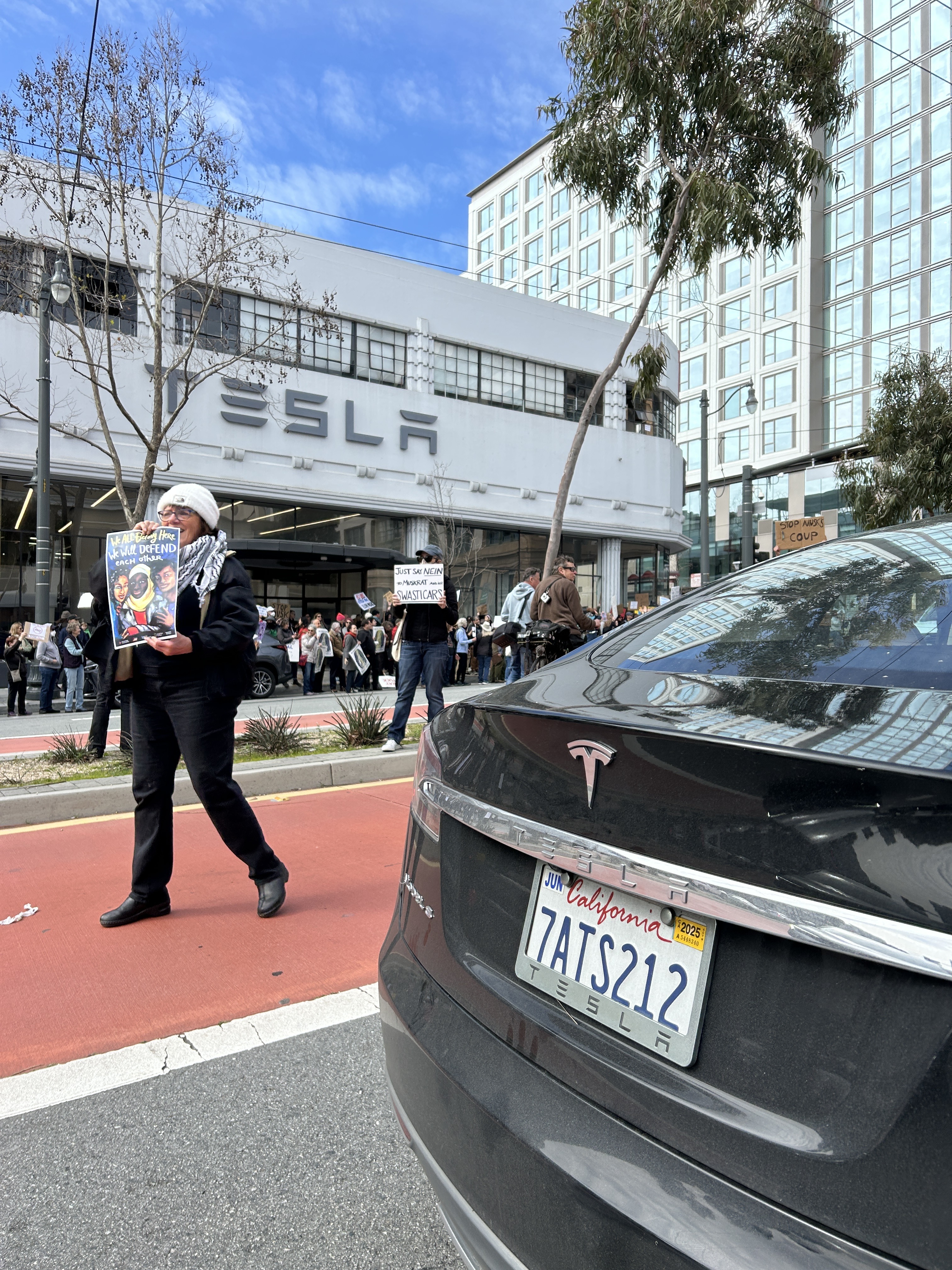 A person holds a protest sign near a Tesla building. A black Tesla with a California license plate is in the foreground. Several people and signs are visible.