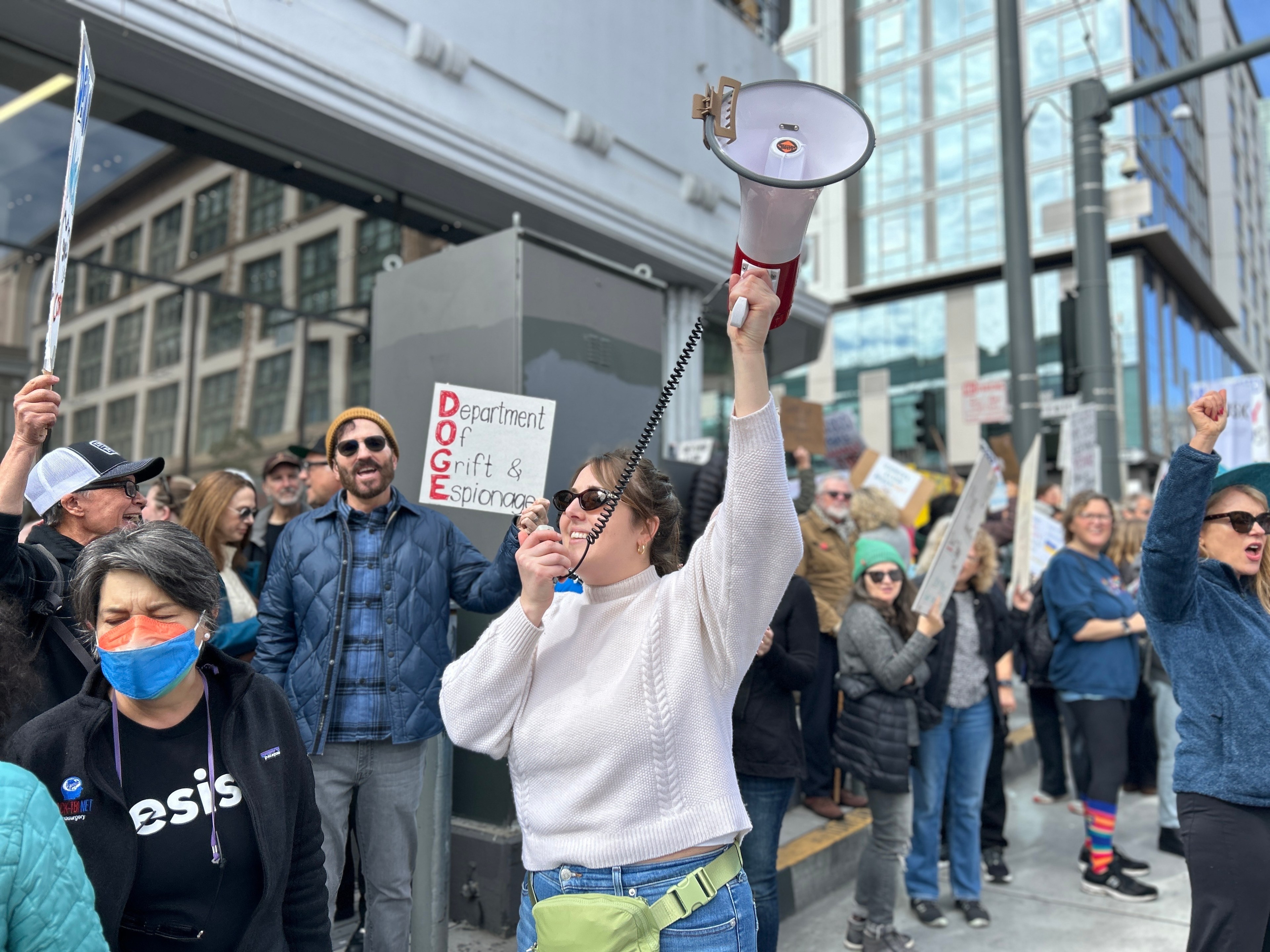 A group of people is gathered in a city street protest. One person holds a megaphone, and another displays a sign reading &quot;DOGE: Department of Grift &amp; Espionage.&quot;