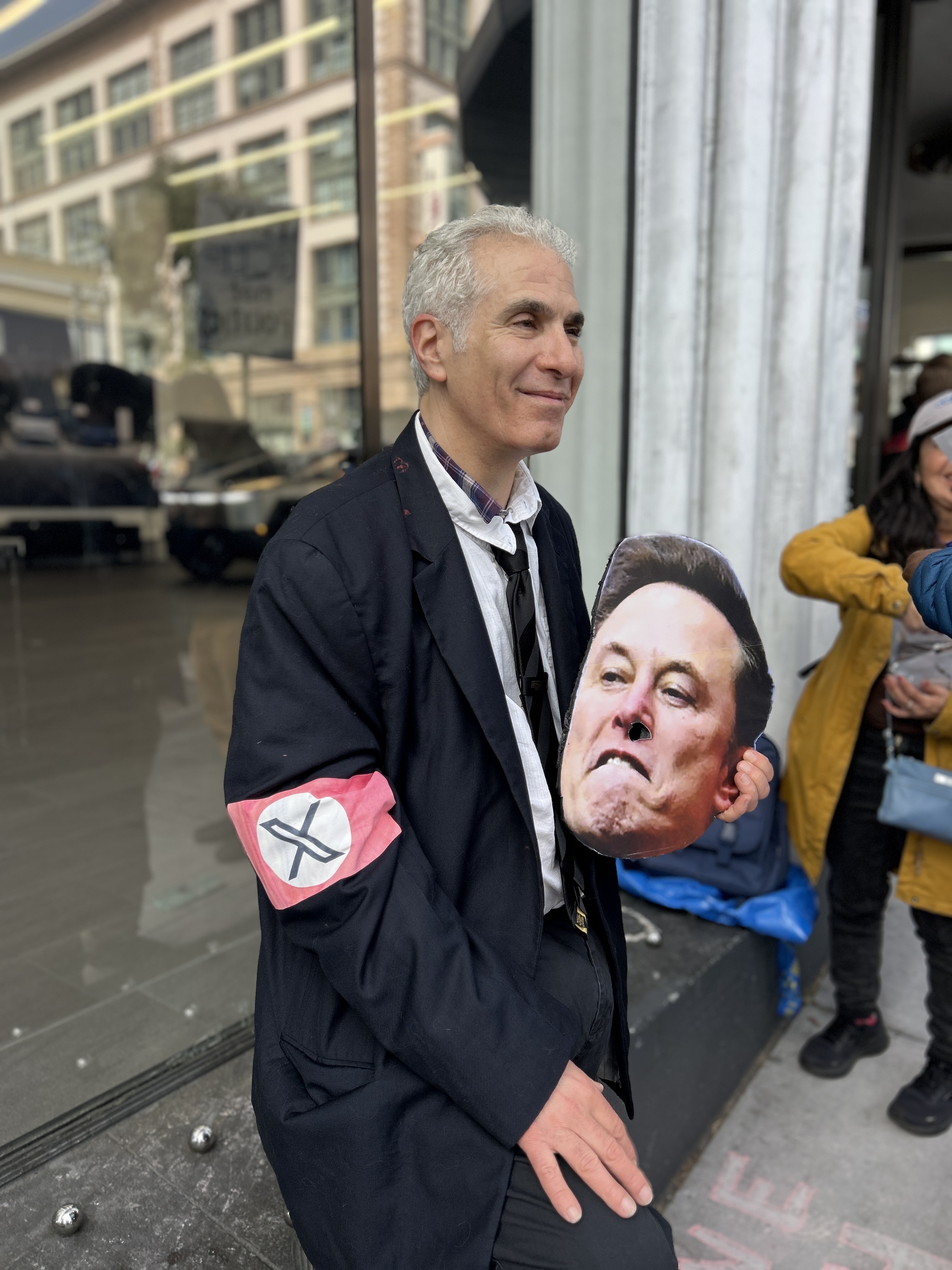 A person with short gray hair stands outside, wearing a dark coat with a red armband featuring an &quot;X.&quot; They're holding a cutout of a man's face.