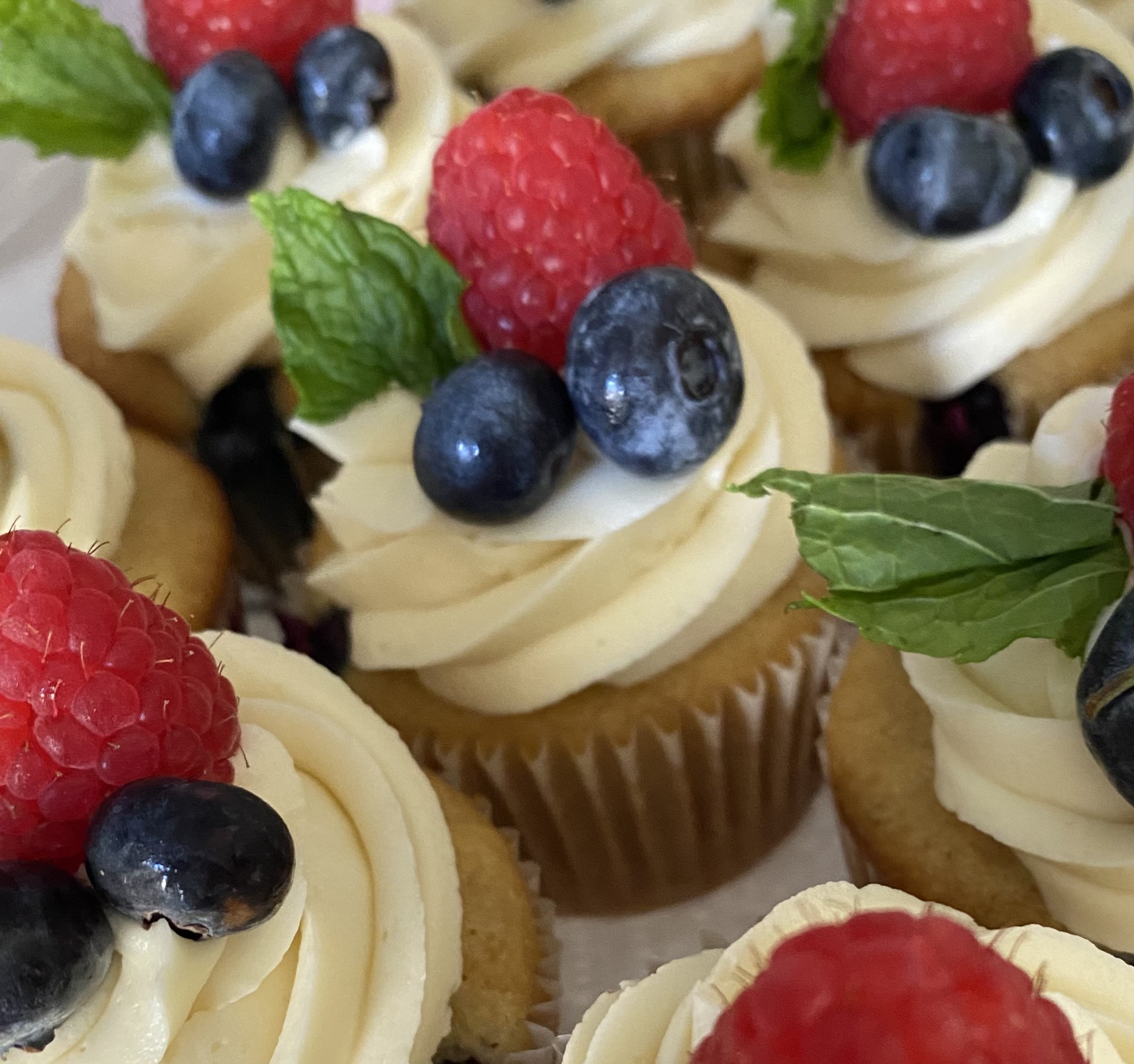 The image shows cupcakes topped with swirls of cream, fresh raspberries, blueberries, and mint leaves arranged attractively.