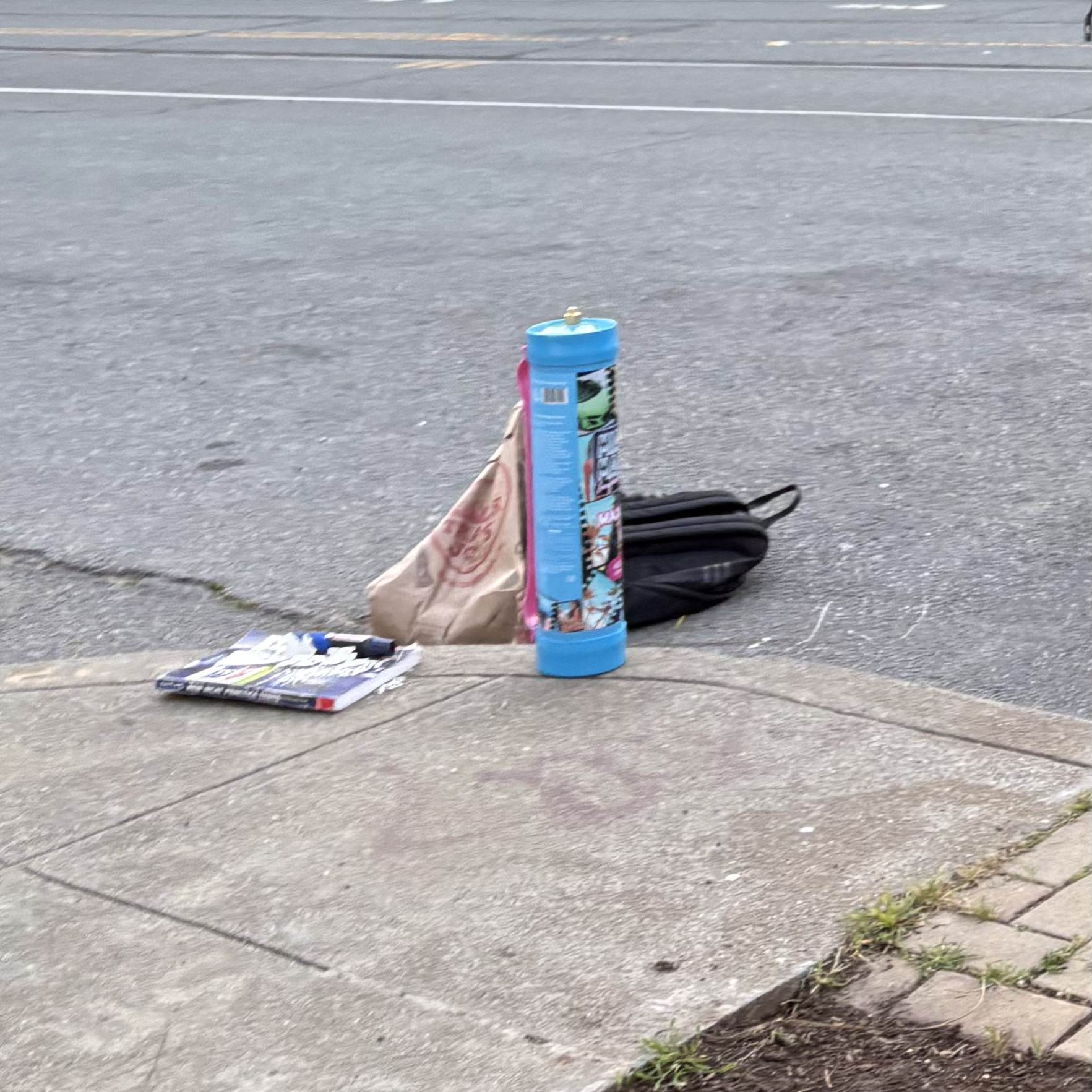 A cylindrical object, paper bag, black bag, and magazine lie on a sidewalk near a street with residential buildings in the background.