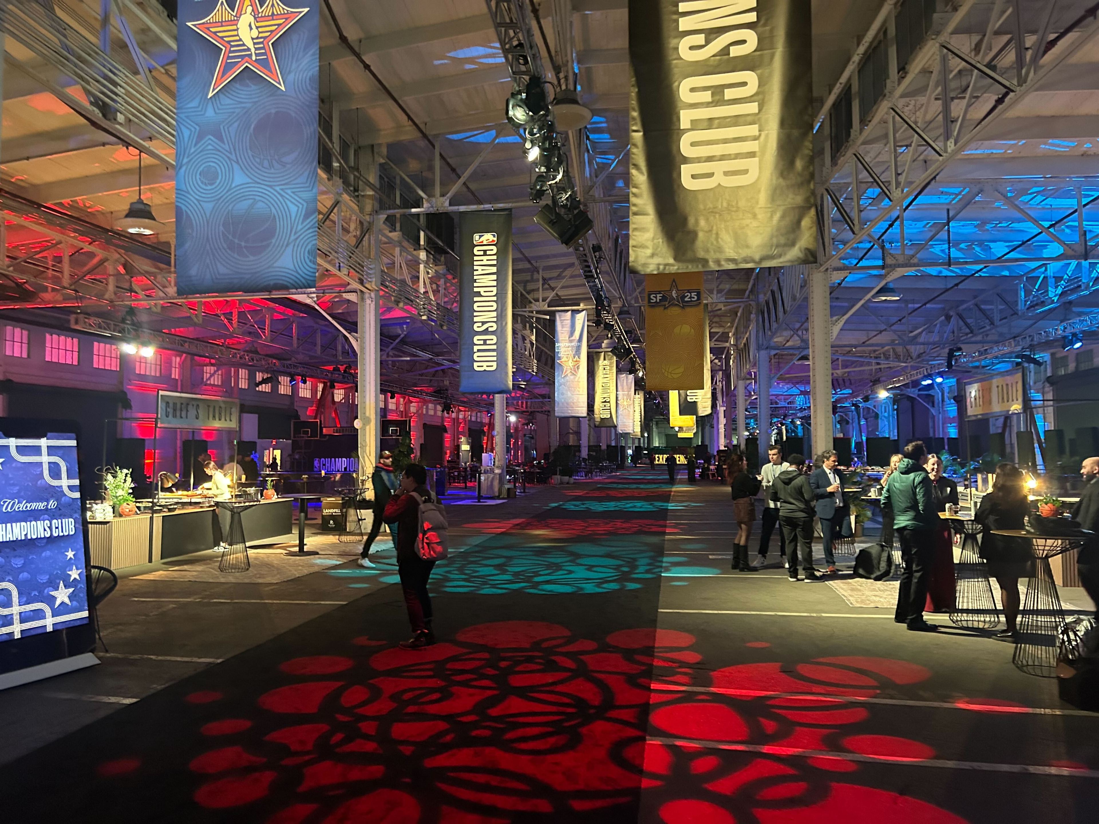 The image shows an indoor event space with colorful lighting, banners reading &quot;Champions Club,&quot; and people mingling around tables. A chef's station is visible.