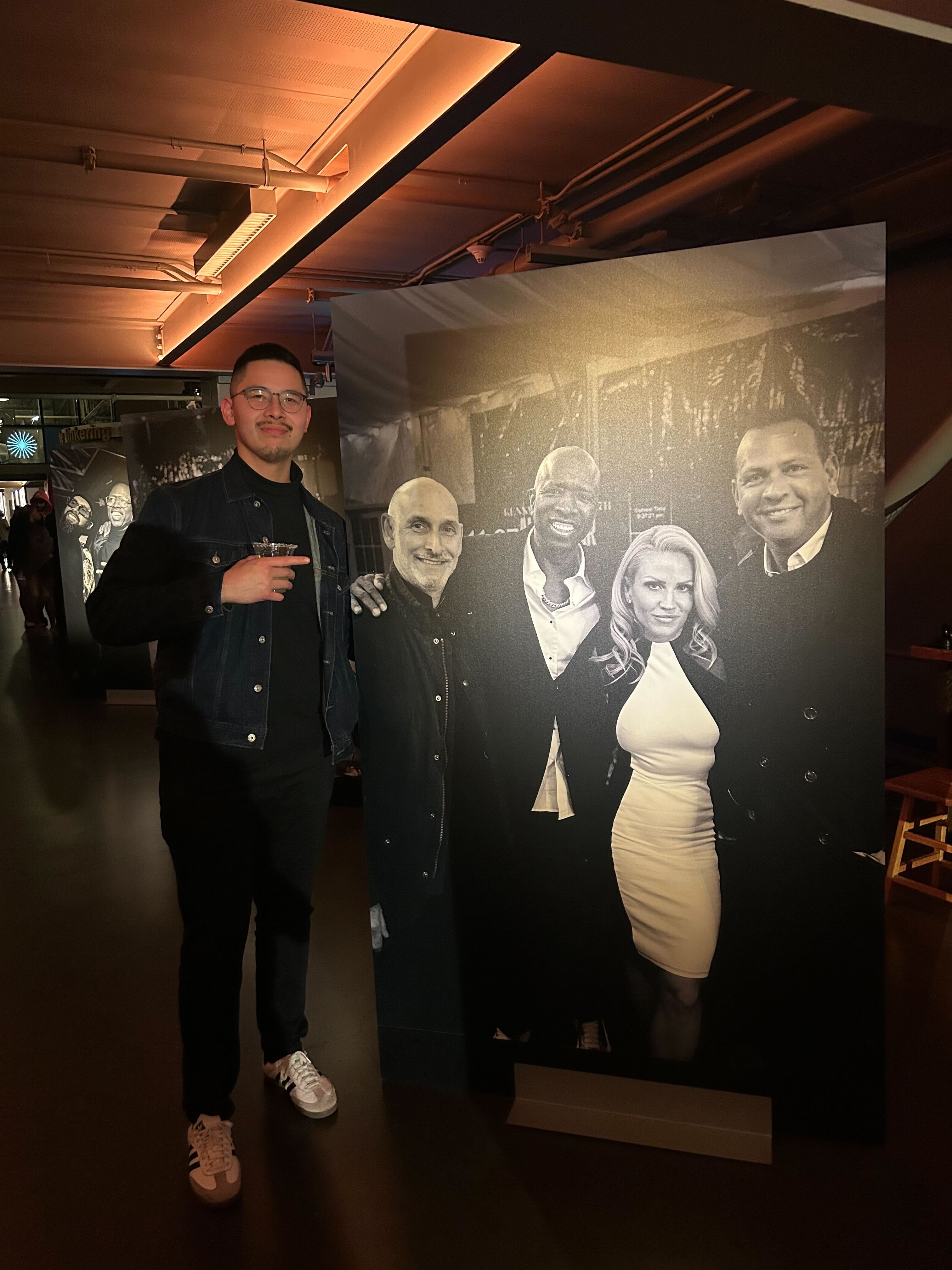 A man stands in a dimly lit room, pointing at a large black-and-white poster featuring four smiling people, two men and a woman, dressed in formal attire.