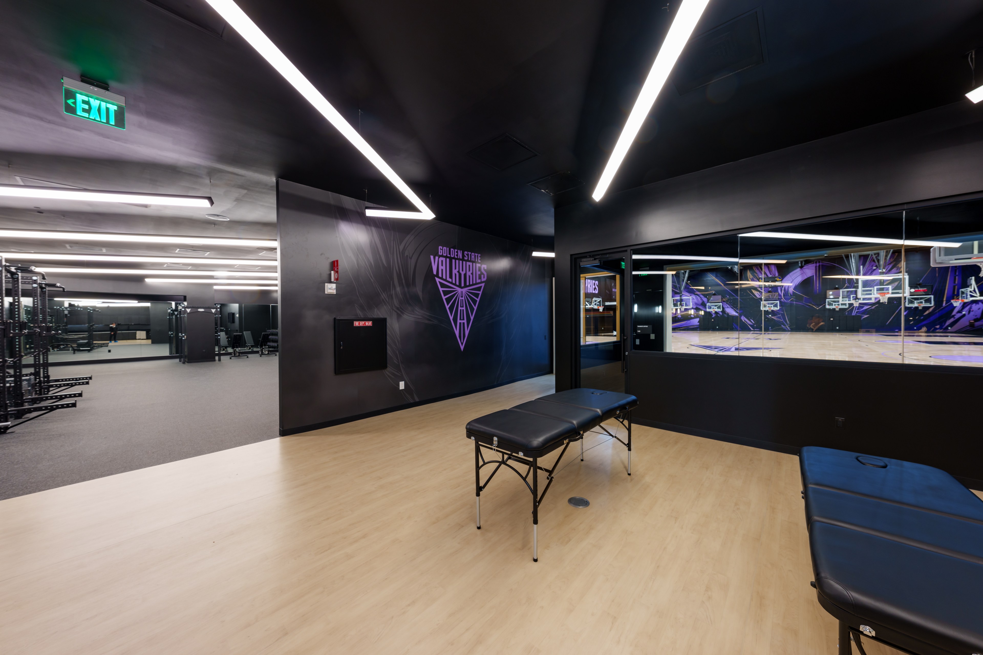 The image shows a sleek sports facility with black walls, purple decor, a training room with massage tables, a weightlifting area, and a view into a basketball court.