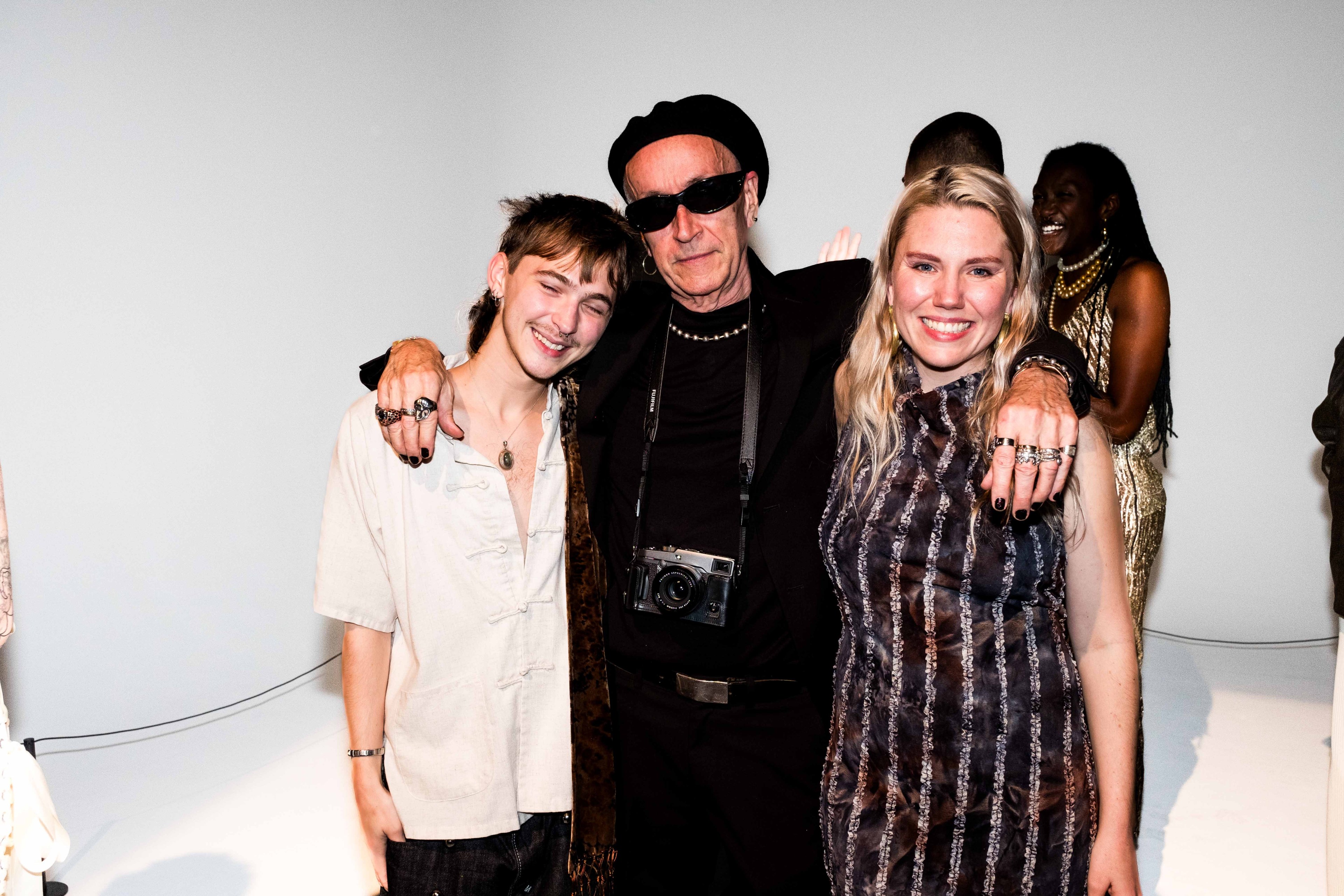 Three people are standing together, smiling. The person in the middle wears black clothing and sunglasses, holding a camera. Others in the background are laughing.