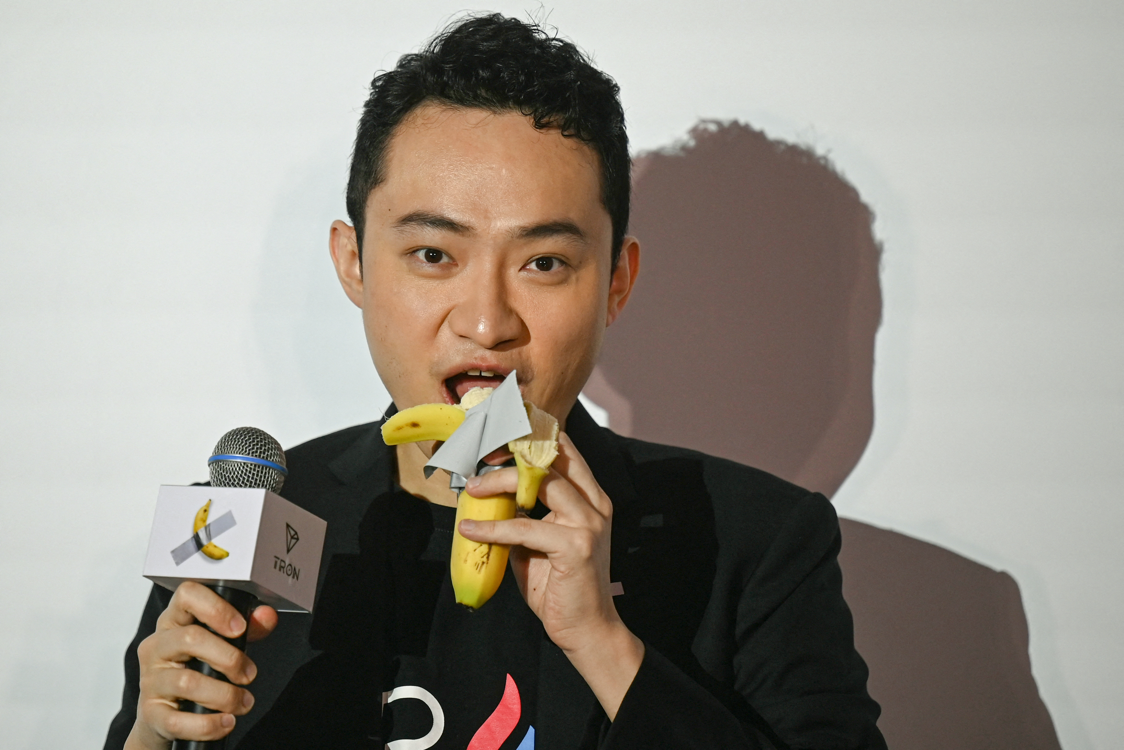 A person holds a microphone with a banana logo and eats a banana wrapped with paper. The background is plain, and the person is wearing black.