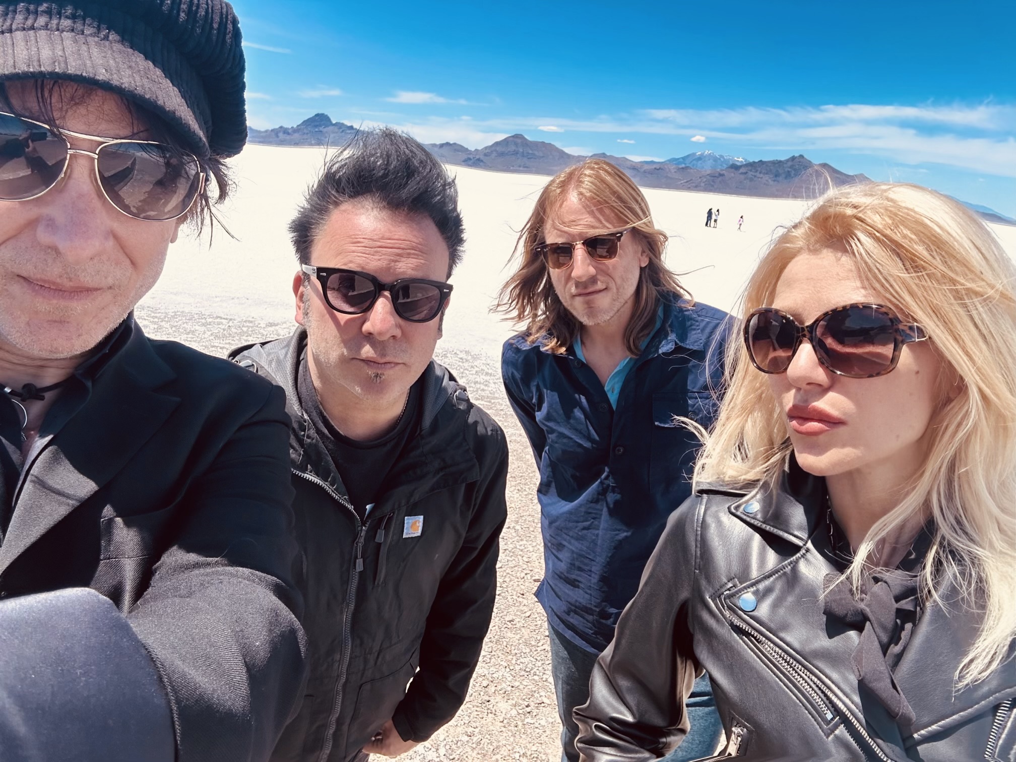 Four people wearing sunglasses are standing together in a sunny, desert-like landscape with mountains in the background. They're all dressed in dark, casual clothing.