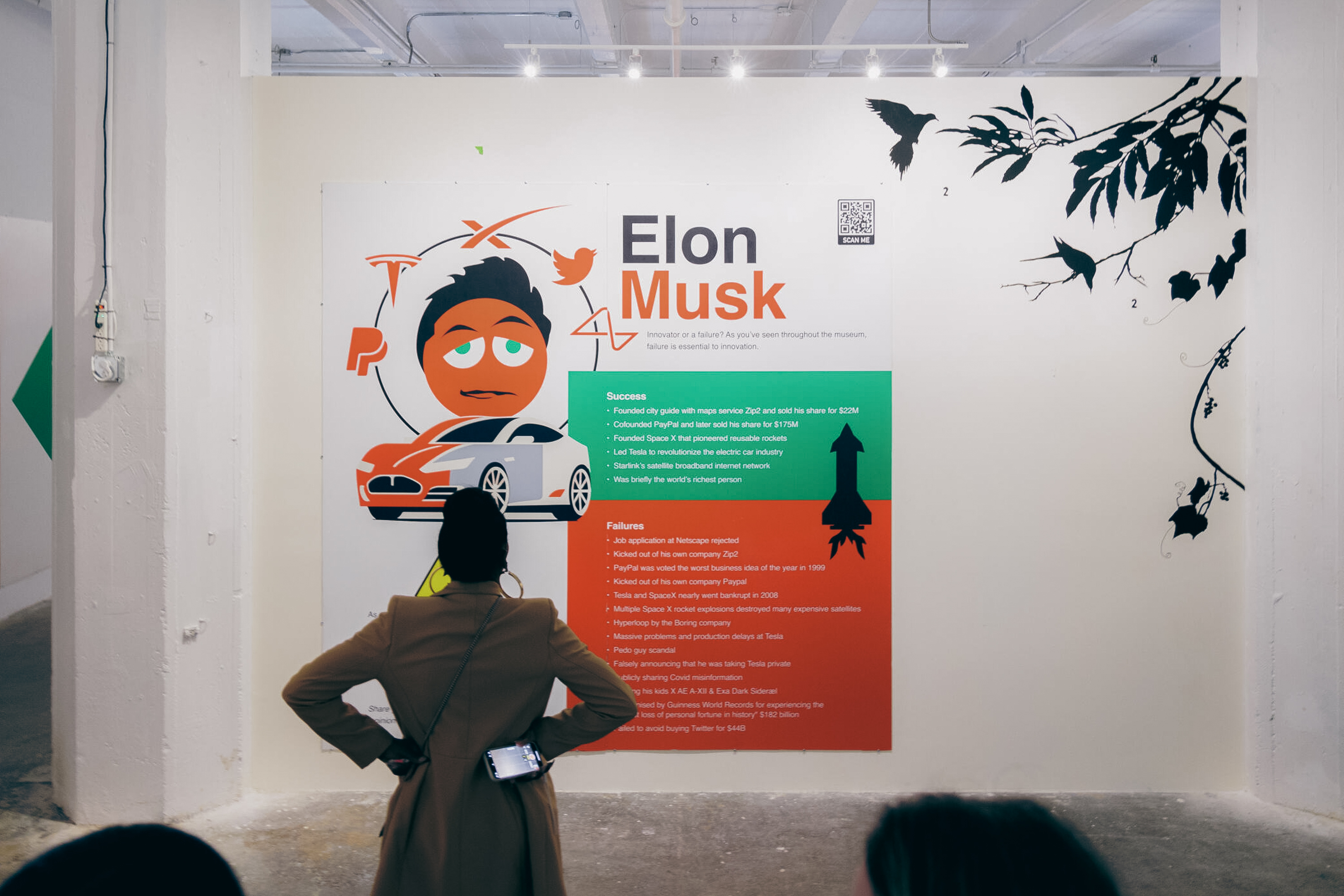 A person stands in front of a wall display featuring an illustration and text about Elon Musk. The display includes both successes and failures, with stylized graphics.