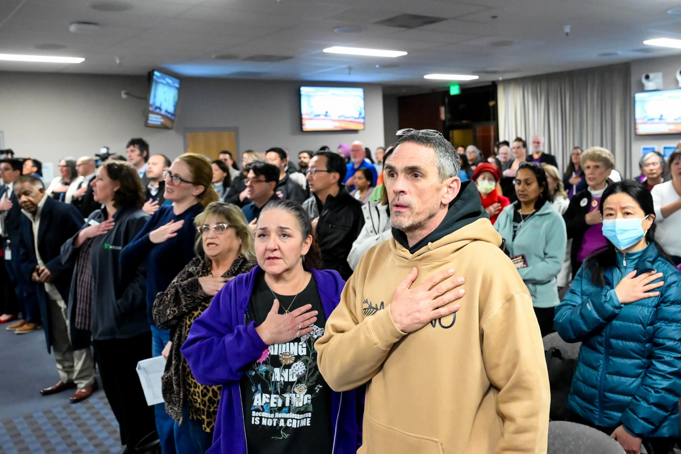 A diverse group of people stands together in a room, each with a hand over their heart, facing forward. Some are wearing masks and appear attentive.