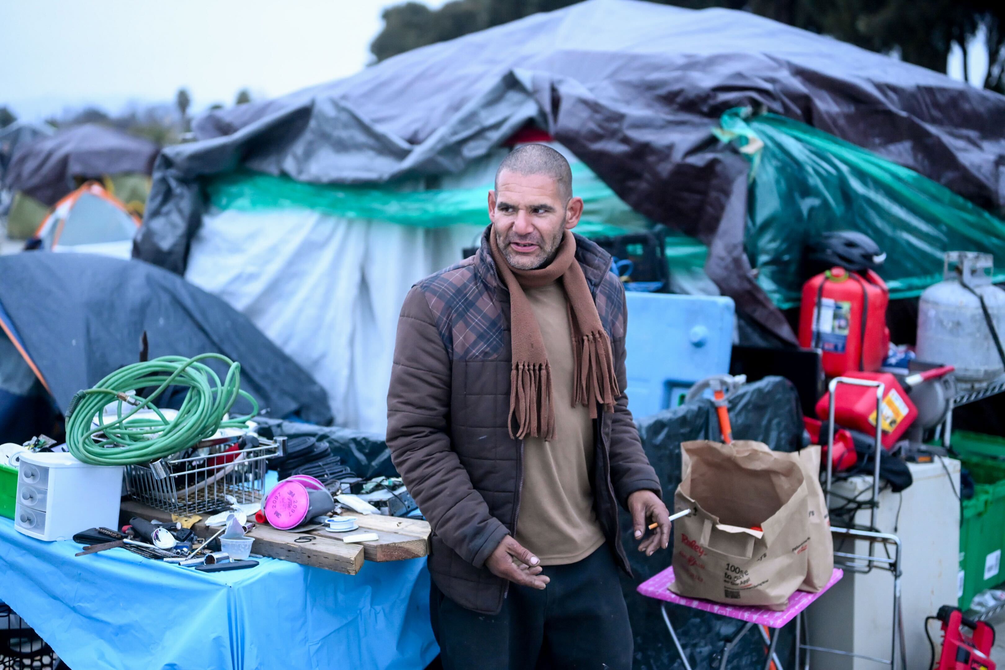 A man stands in front of a makeshift shelter with tarps. There's a table cluttered with various items like wires, tools, and a paper bag.