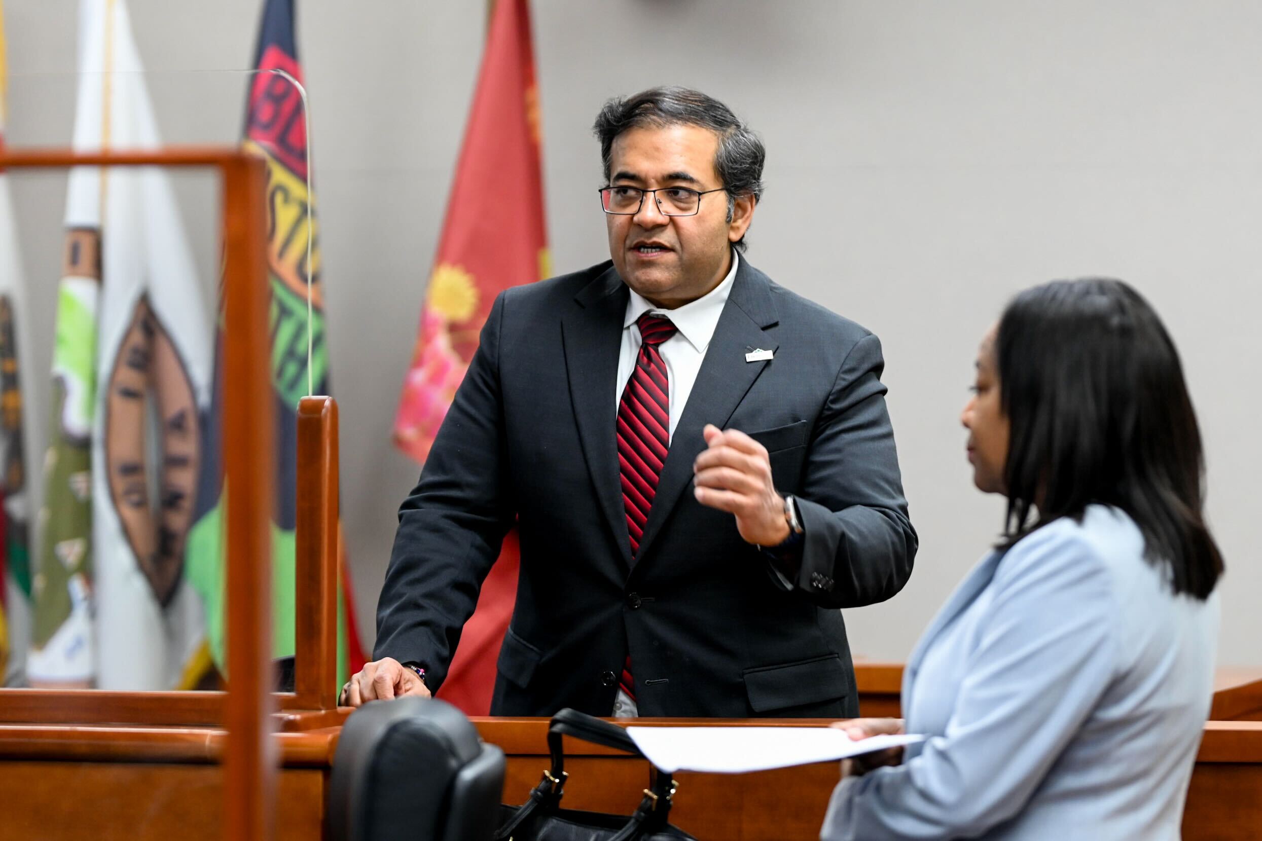 A man in a suit and red tie speaks in a courtroom setting, gesturing with his hand. A woman in a light-colored outfit holds papers, with flags visible in the background.