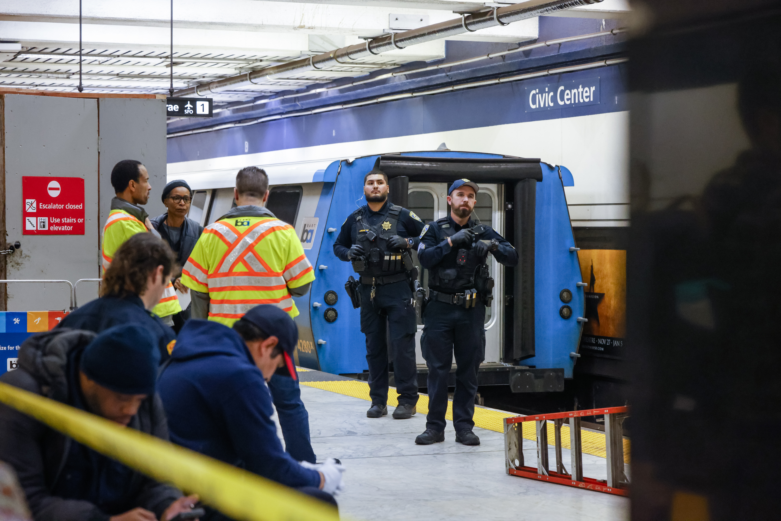 Two police officers stand near a train at Civic Center station. Workers in high-visibility jackets converse nearby, with a few people seated in the foreground.