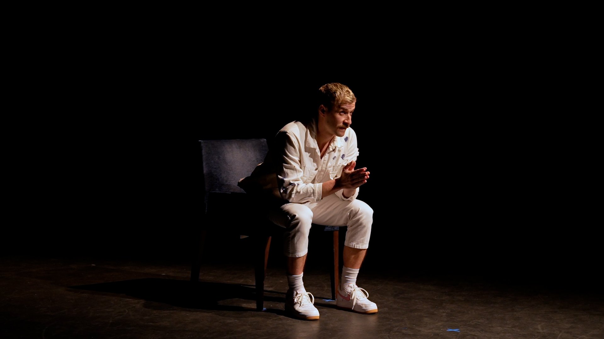 A person in white clothing sits on a dark chair against a black background, leaning forward with hands clasped, appearing thoughtful or engaged.