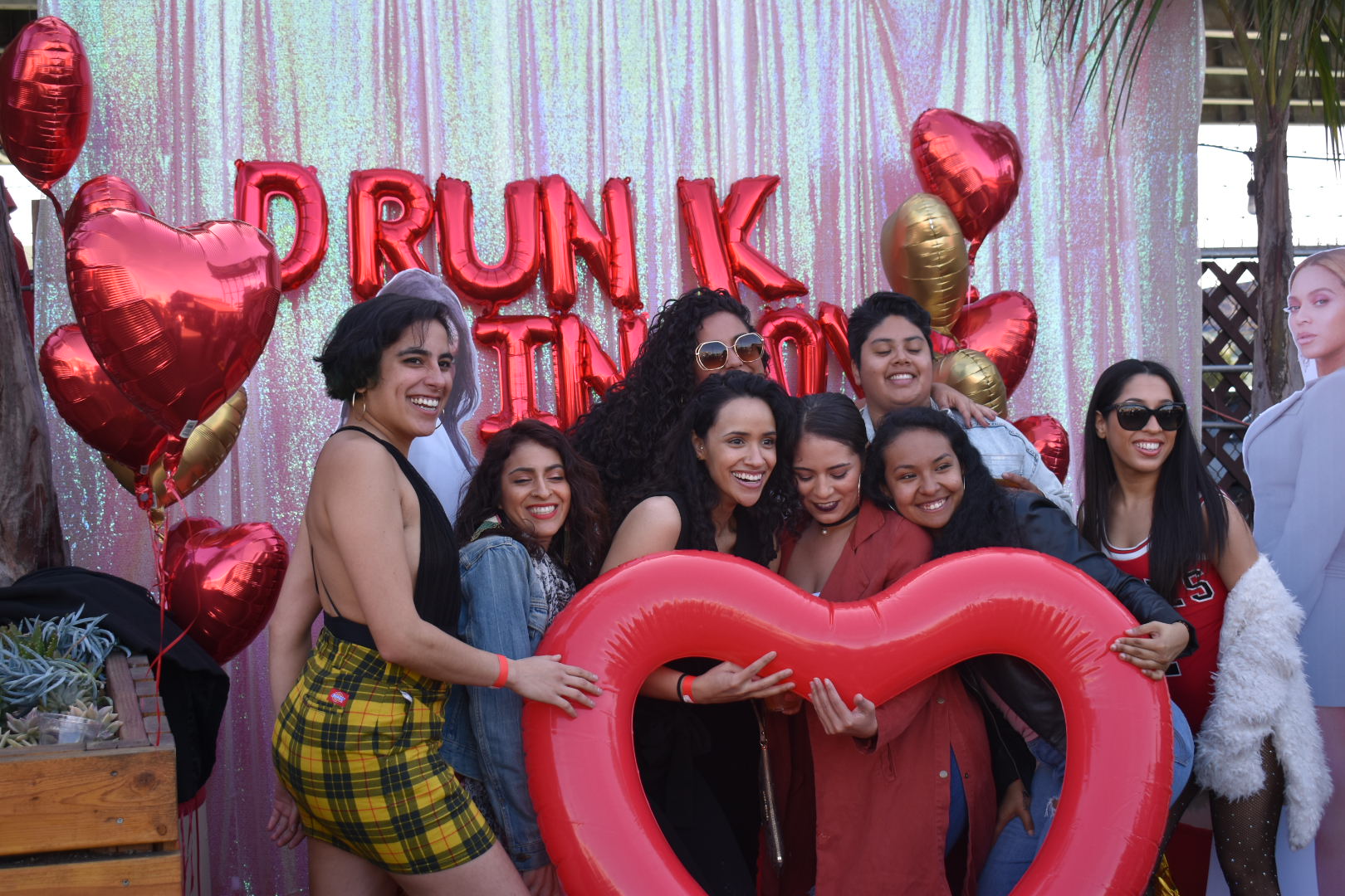 A group of smiling people pose in front of a shiny backdrop with red heart balloons. They hold a large inflatable red heart and appear joyful.