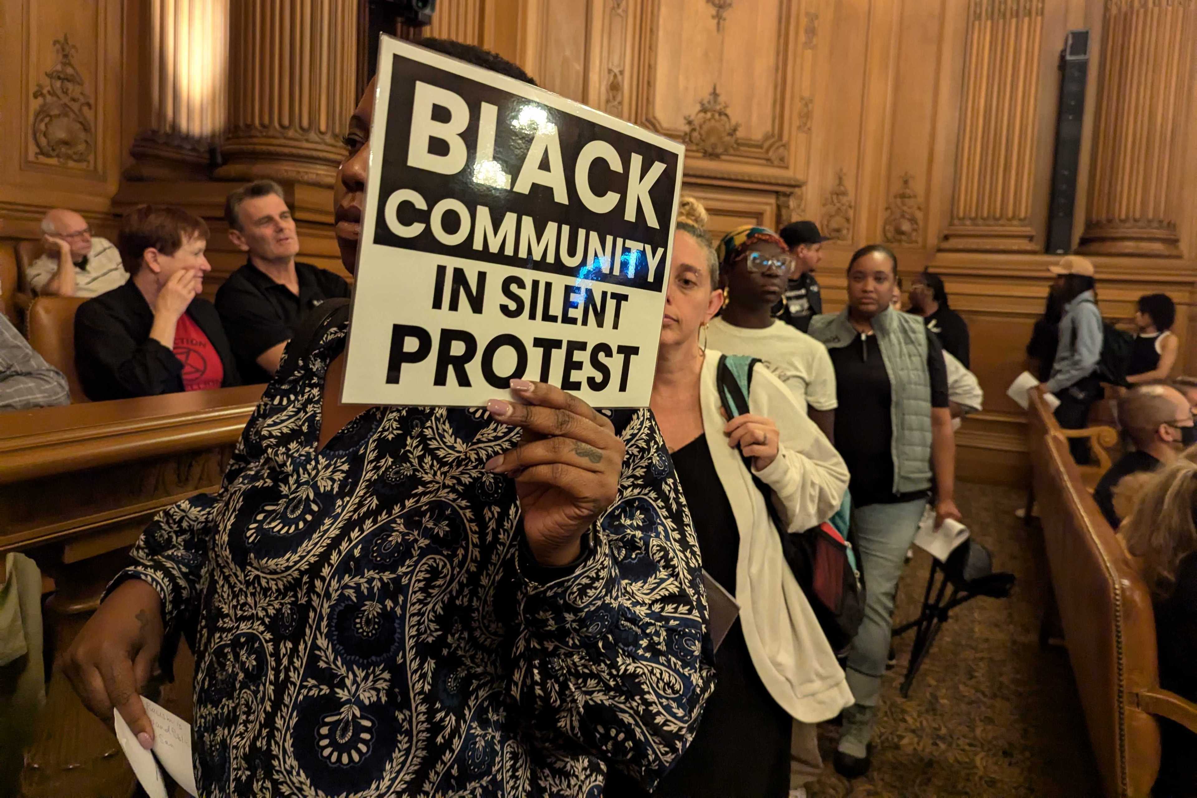 A person holds a sign reading "Black Community in Silent Protest" in a wooden-paneled room filled with people, suggesting a gathering or meeting.
