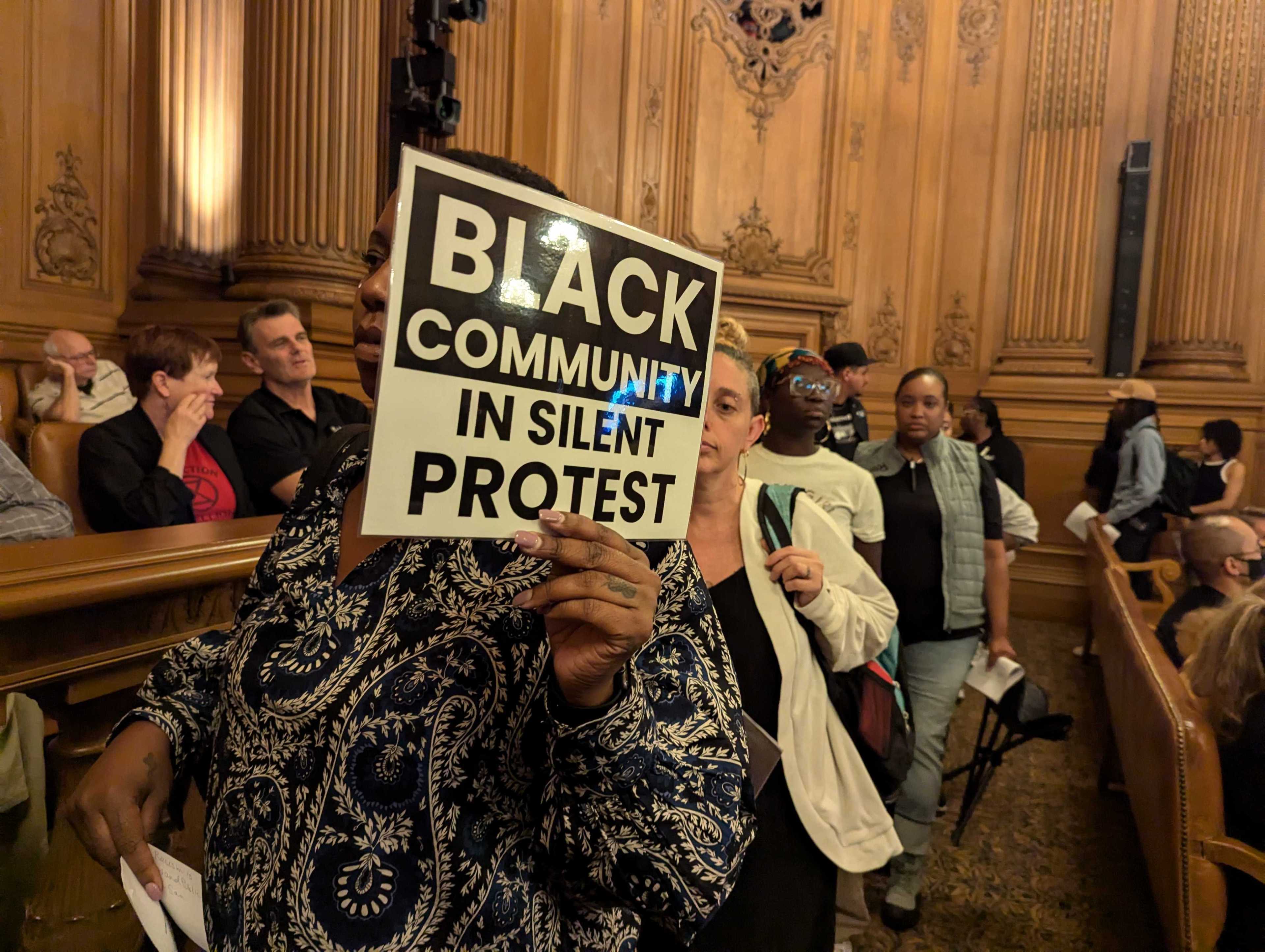 A person holds a sign reading "Black Community in Silent Protest" in a wooden-paneled room filled with people, suggesting a gathering or meeting.