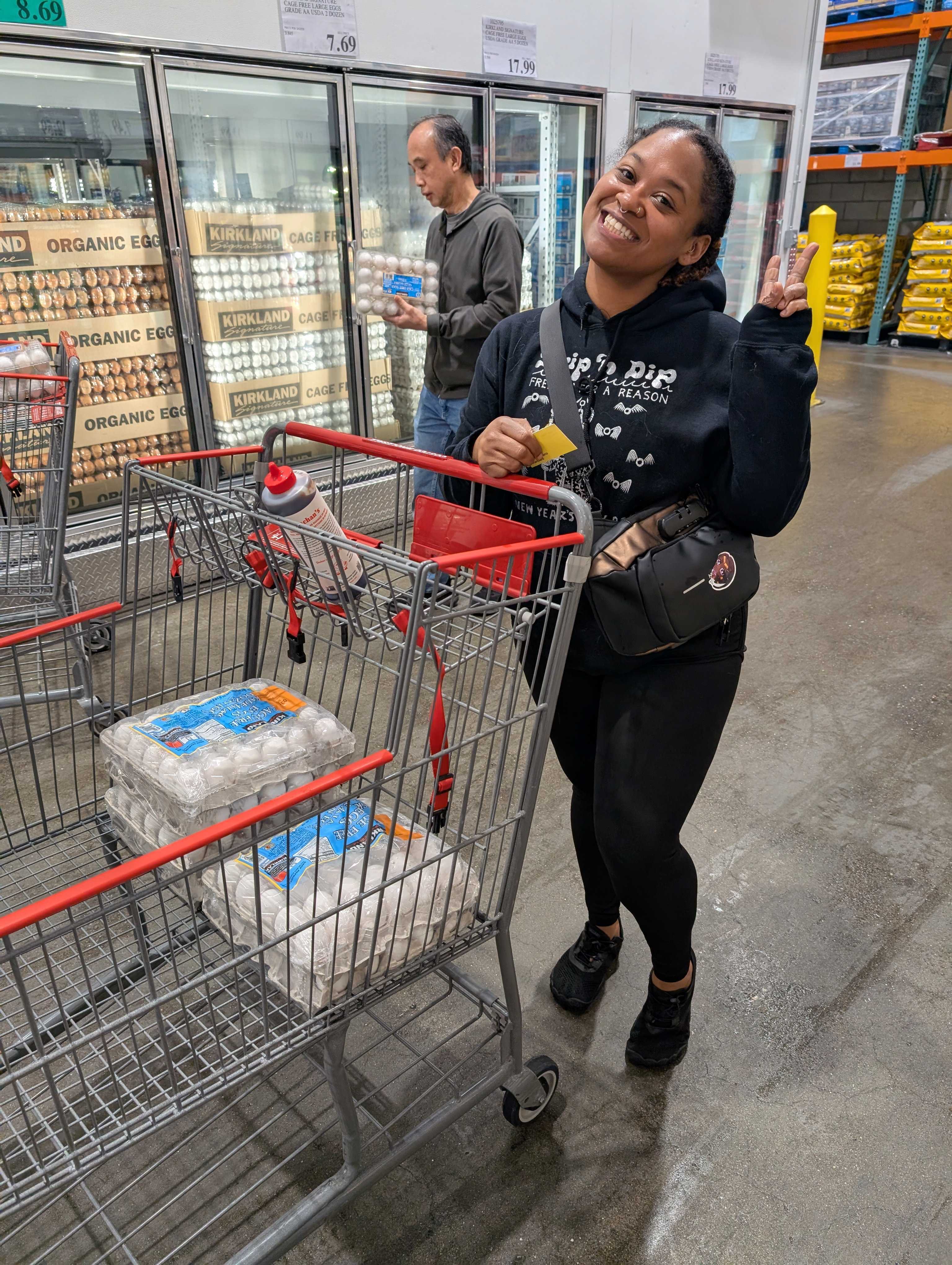 A woman smiles and shows a peace sign next to a shopping cart with eggs. Behind her, a man selects eggs from a refrigerated display in a store.