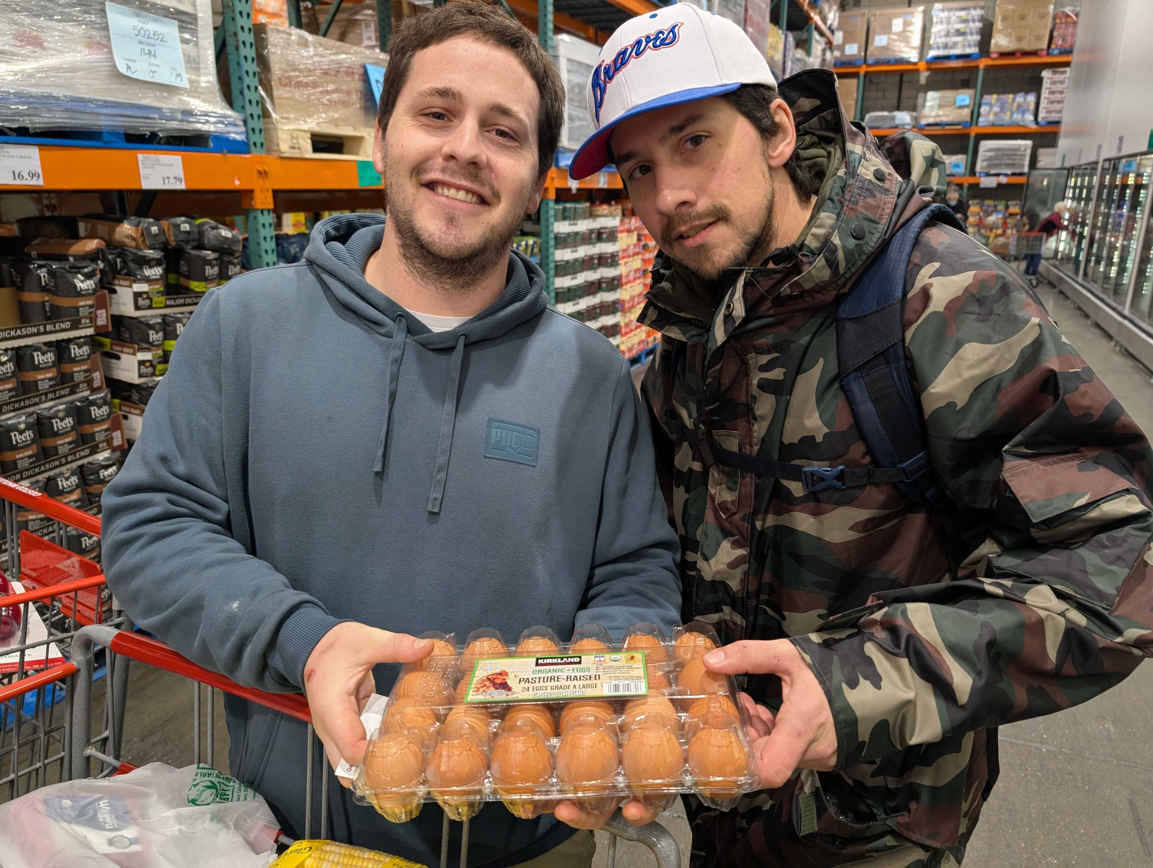 Two people are in a store, both smiling and holding a carton of eggs. One is wearing a blue hoodie; the other has a camouflage jacket and a white baseball cap.