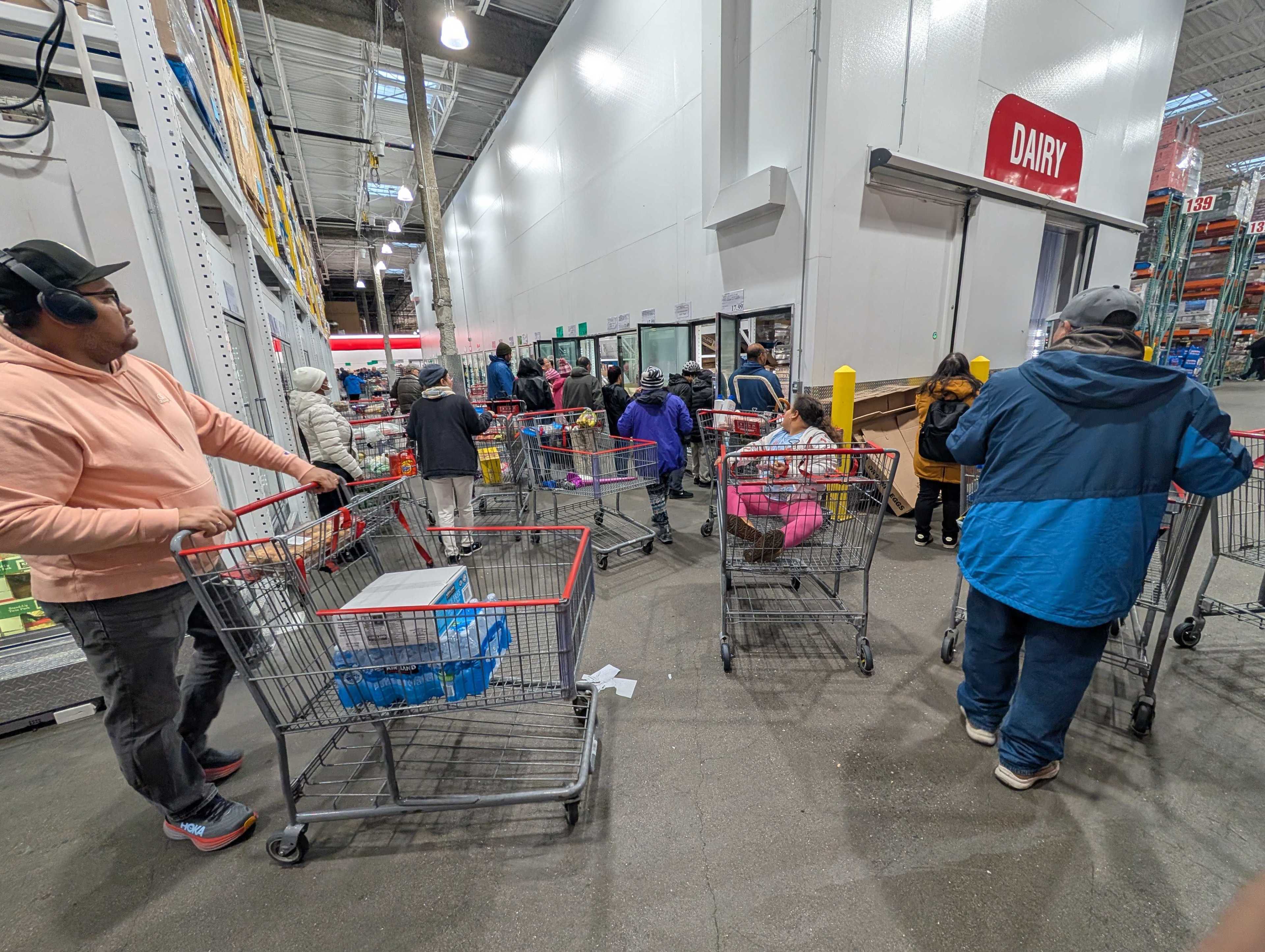 People with shopping carts are lining up in a large store aisle under a sign labeled &quot;Dairy.&quot; They are browsing shelves and refrigerators.