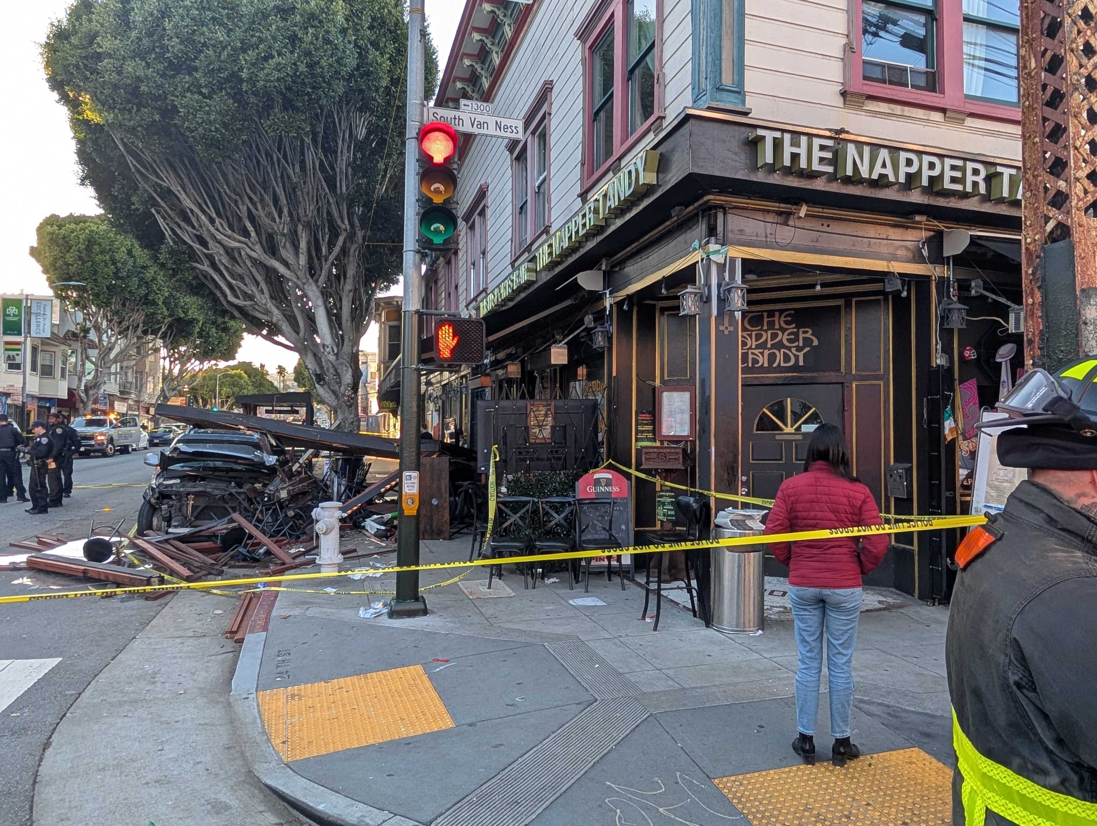 A car crash scene outside "The Napper Tandy" with debris scattered and caution tape blocking access. Police officers and bystanders are present near the damaged vehicle.