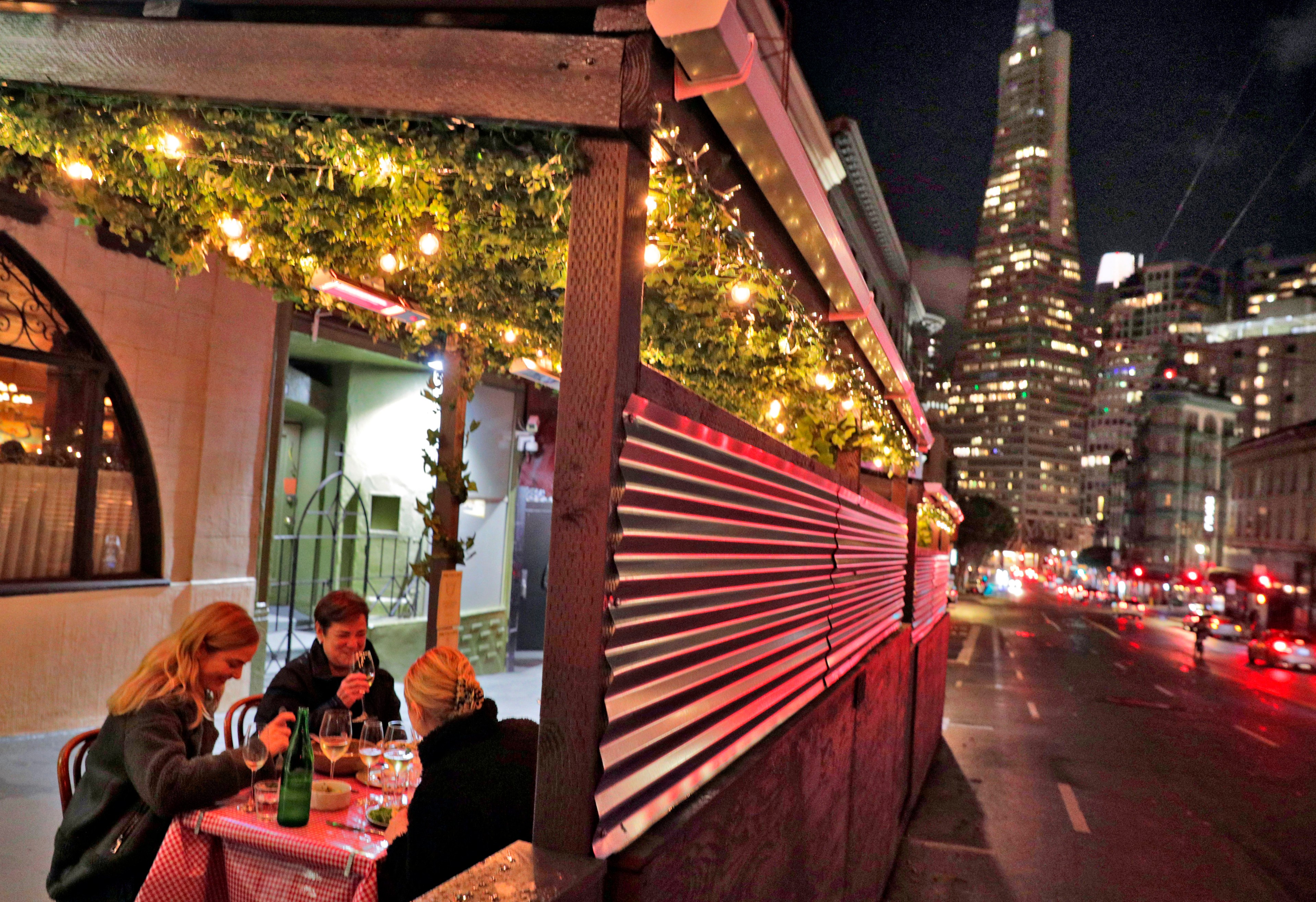 Three people are dining outside under string lights at a table with a checkered cloth. A city street and a tall illuminated building are visible at night.