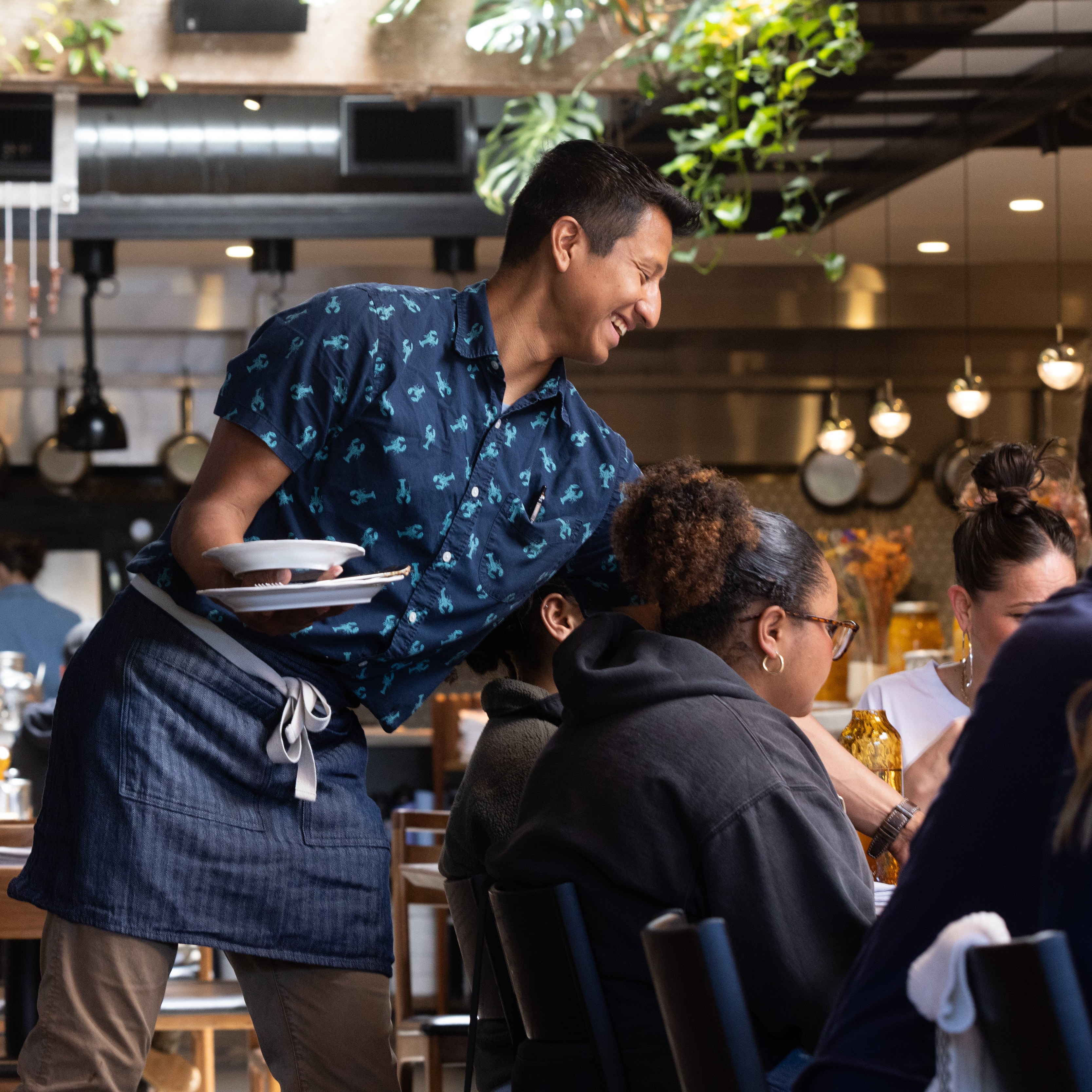 A waiter in a patterned shirt and apron smiles as he serves food to a group of people seated in a lively restaurant with hanging plants and a busy kitchen.