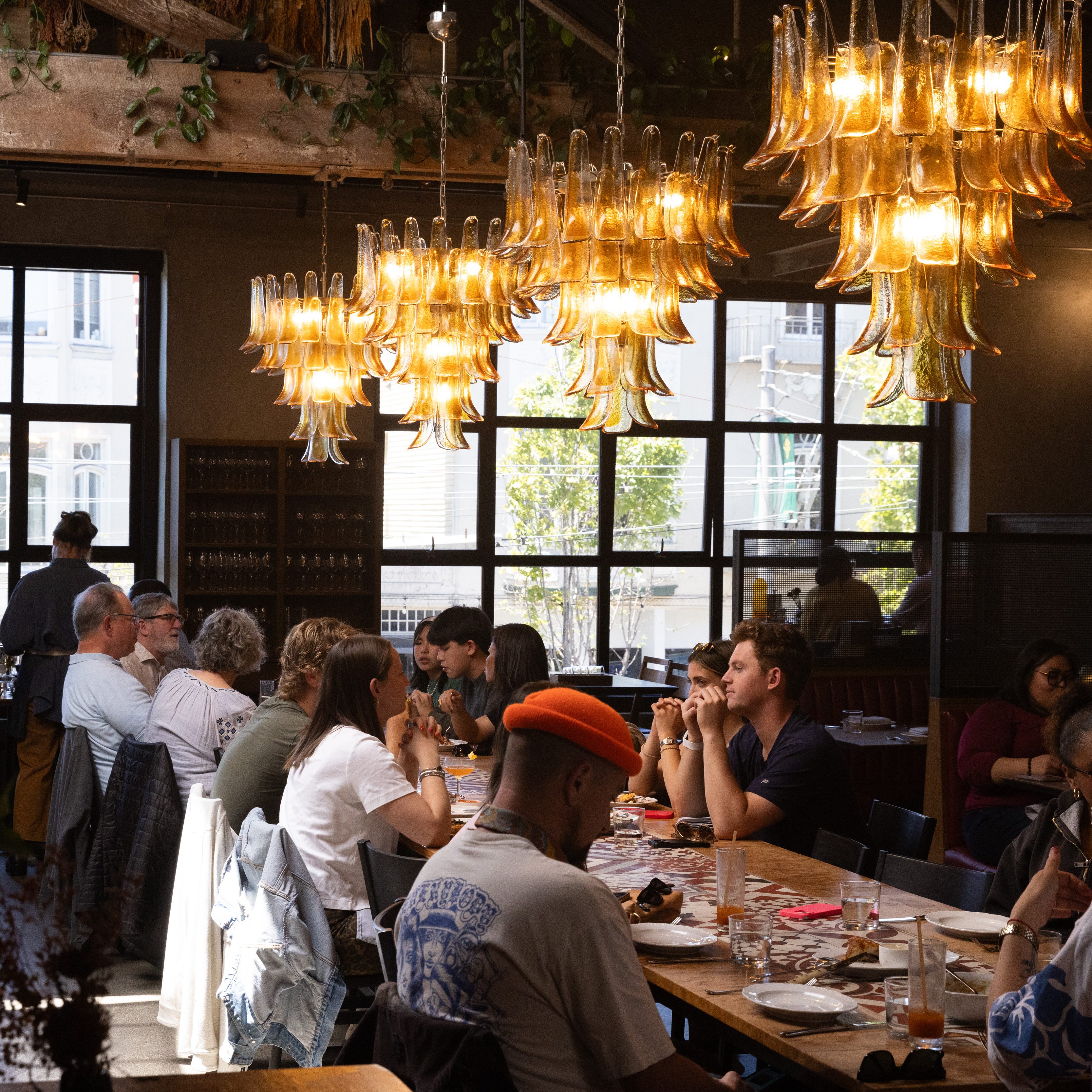 A group of people sit around a long wooden table in a restaurant. Warm light from ornate chandeliers illuminates the scene, creating a cozy ambiance.