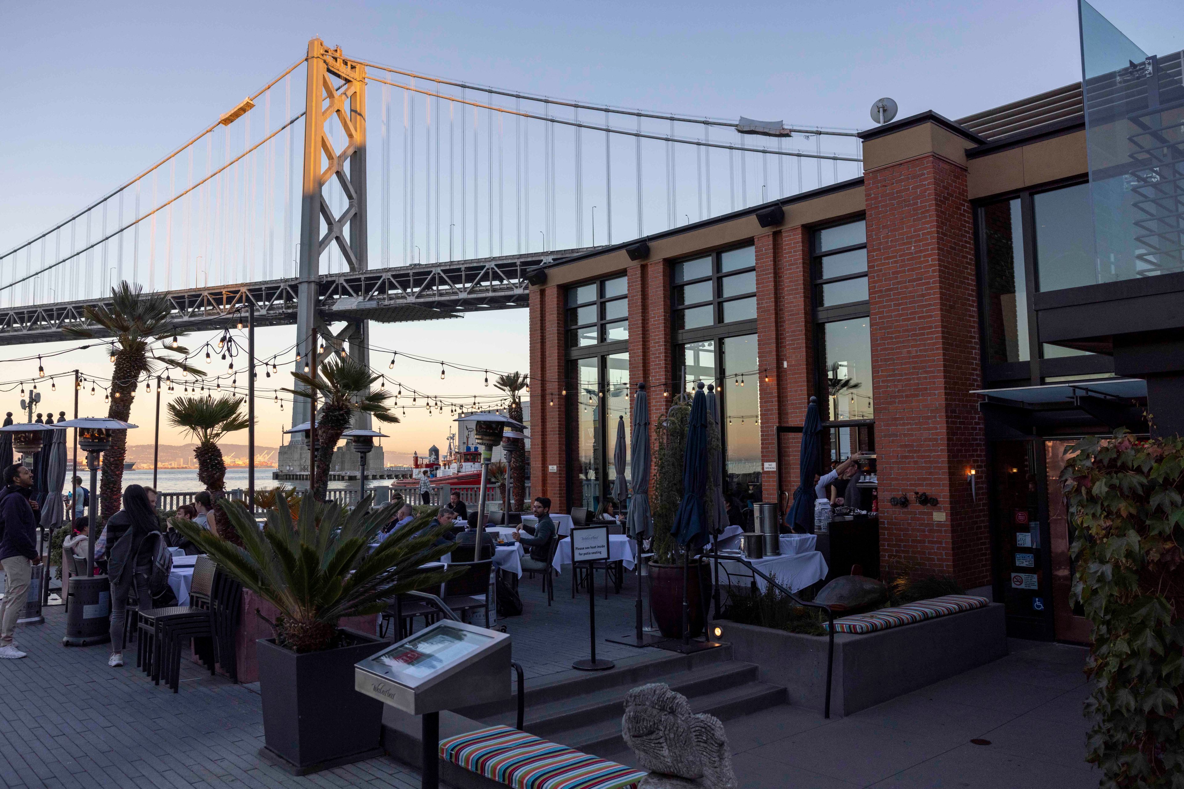 A vibrant outdoor restaurant by the waterfront features palm trees and string lights. A towering bridge spans the background. Diners sit at tables enjoying the view.