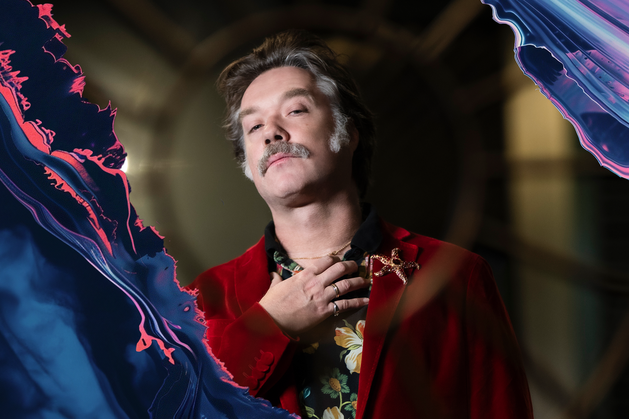 A man with short hair and a mustache wears a red jacket and patterned shirt, holding a hand to his chest. Vivid abstract colors frame the image's sides.