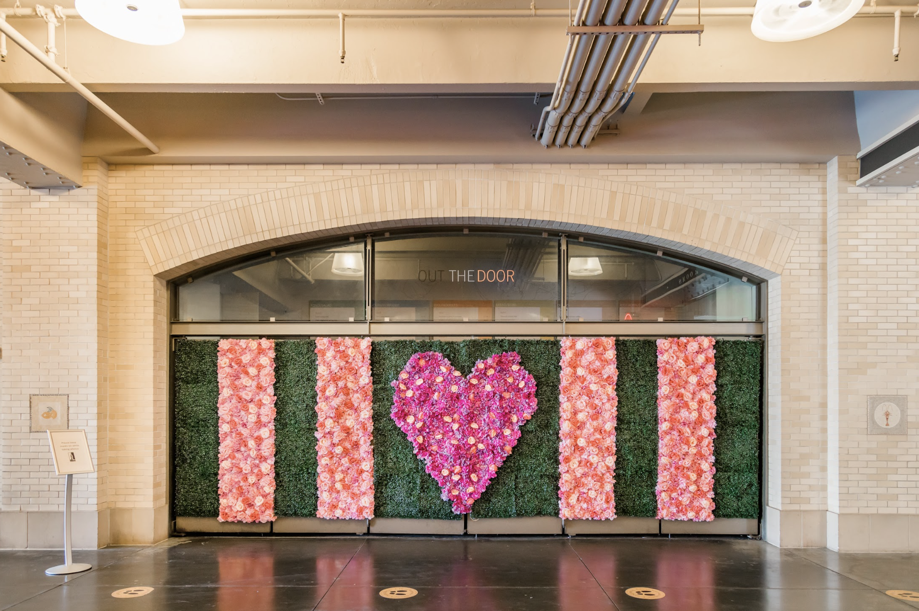 A floral mural features vertical stripes of pink flowers on green foliage, with a purple heart in the center. The backdrop is an arched, glass door.