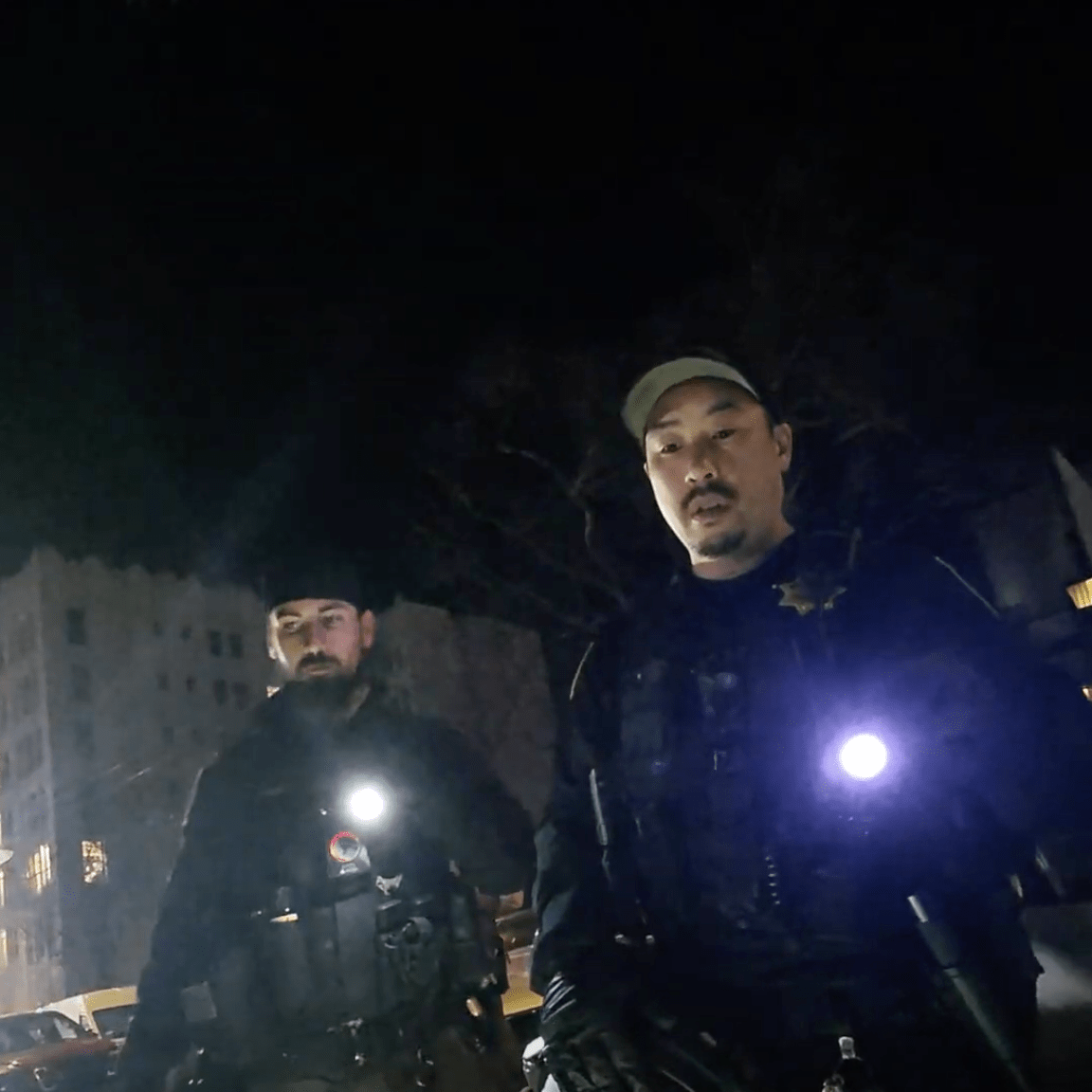 Two officers stand outdoors at night, holding flashlights. They wear tactical gear and appear to be on a street. A building is faintly visible in the background.