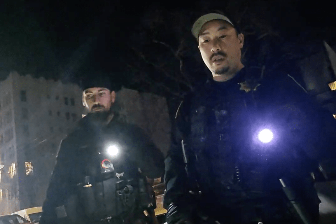 Two officers stand outdoors at night, holding flashlights. They wear tactical gear and appear to be on a street. A building is faintly visible in the background.