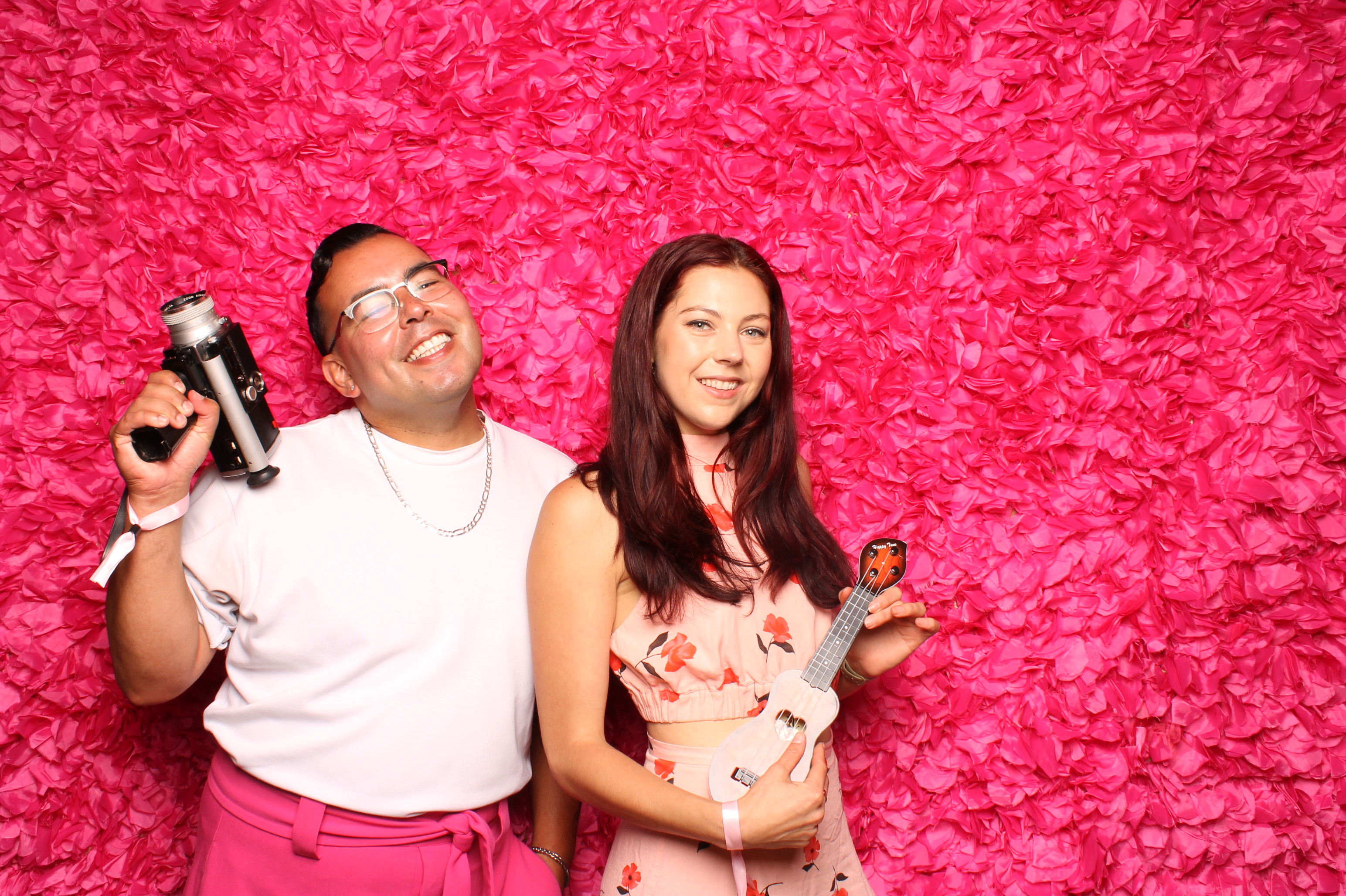 Two people stand in front of a vibrant pink flower wall. One holds a camera and smiles, while the other, wearing a floral outfit, holds a small ukulele.