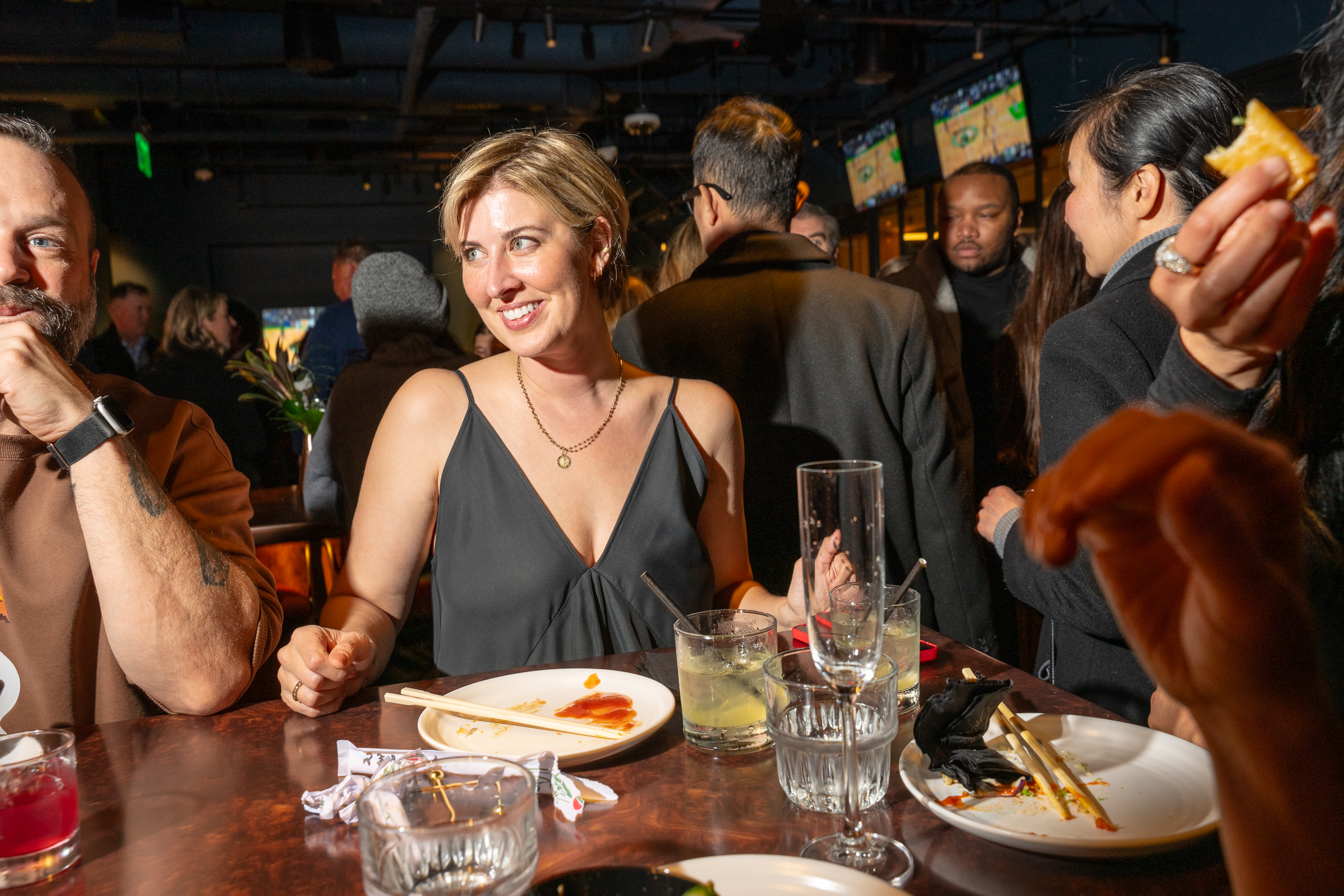 A group of people are at a lively restaurant or bar. A smiling woman in a black dress and short hair sits at a table with drinks and empty plates, surrounded by others.