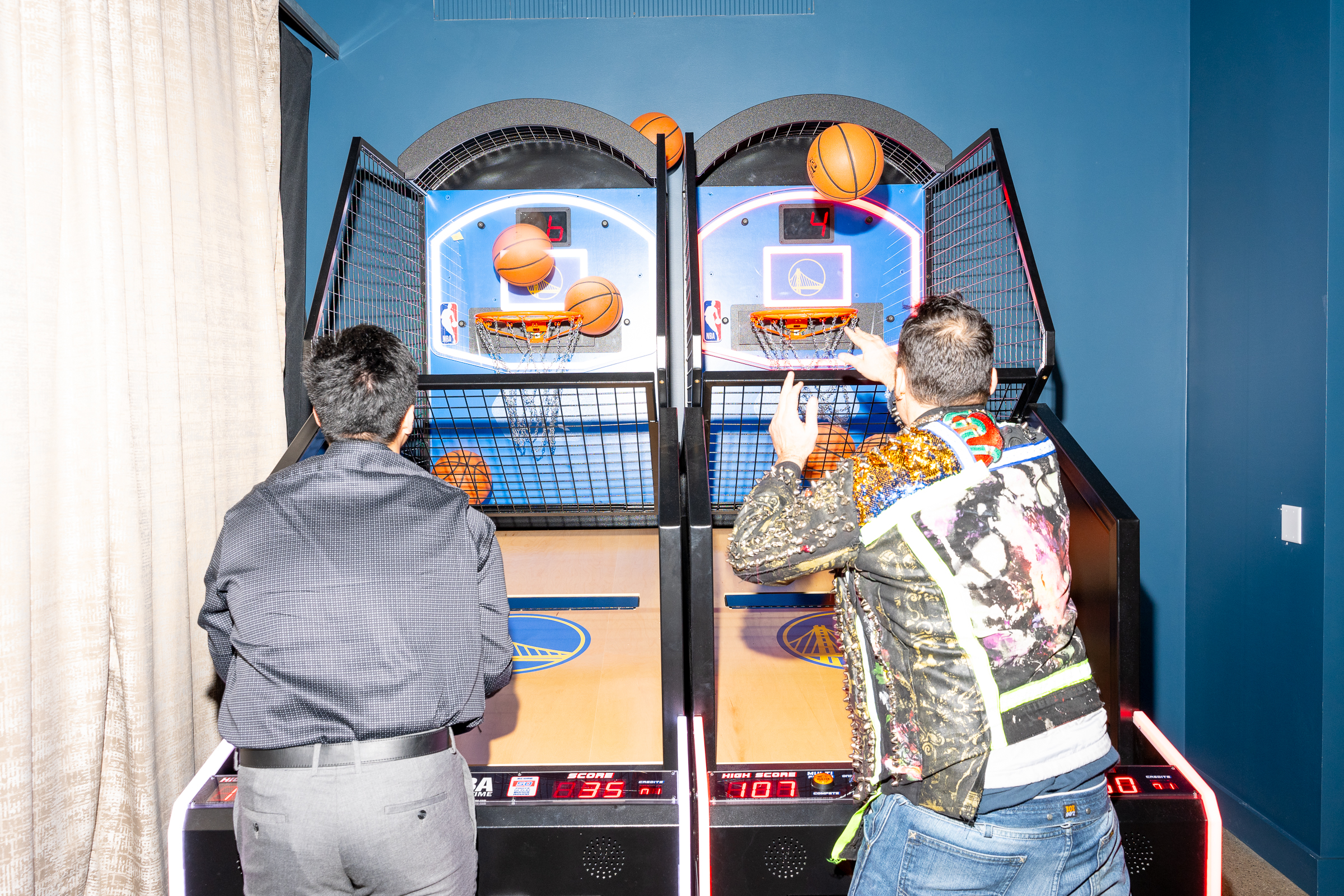 Two people are playing an arcade basketball game. They're focused on shooting basketballs into the hoops, with scores visible below.