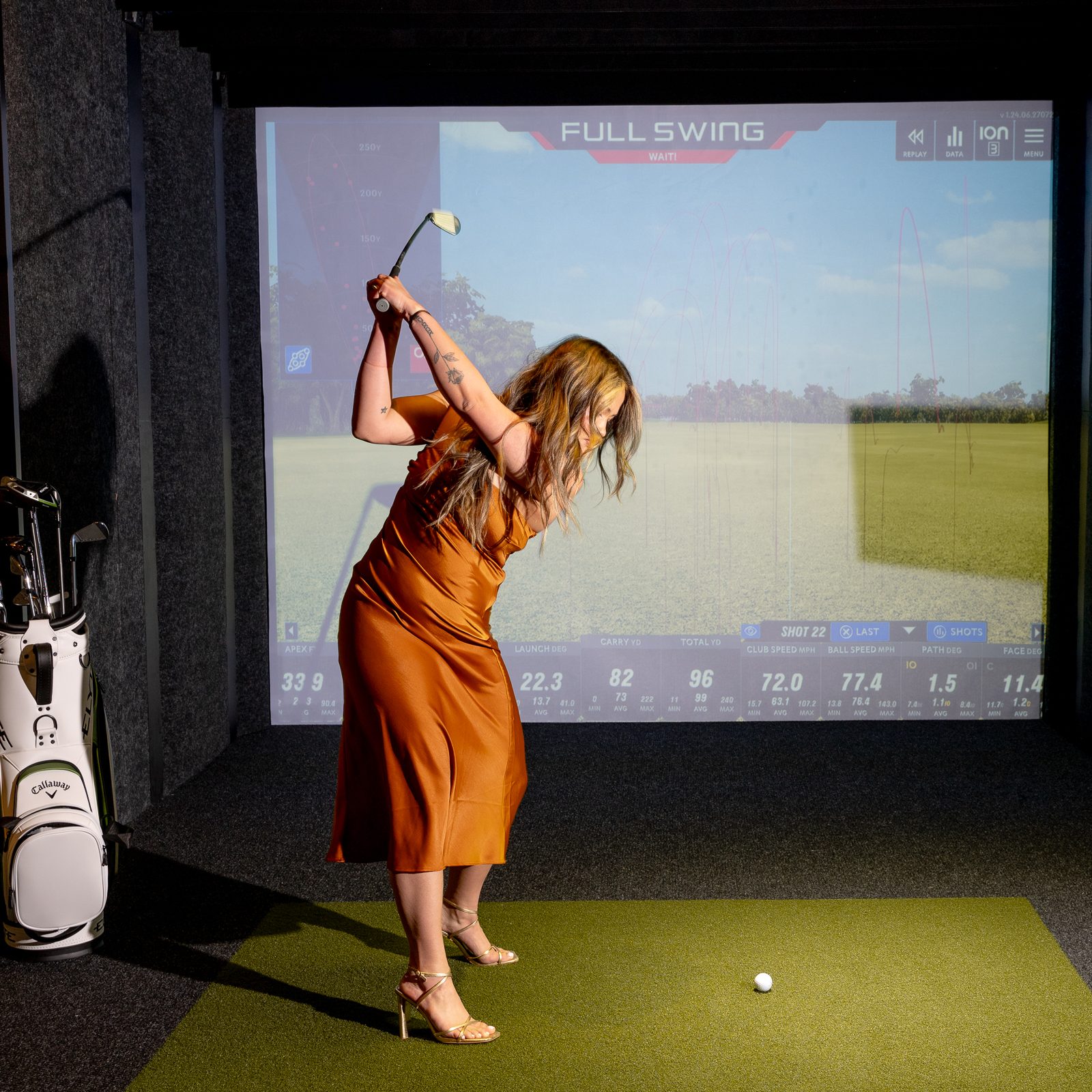 A woman in a satin dress and heels is golfing inside on a simulator, holding a club mid-swing. A bag of golf clubs is visible nearby.