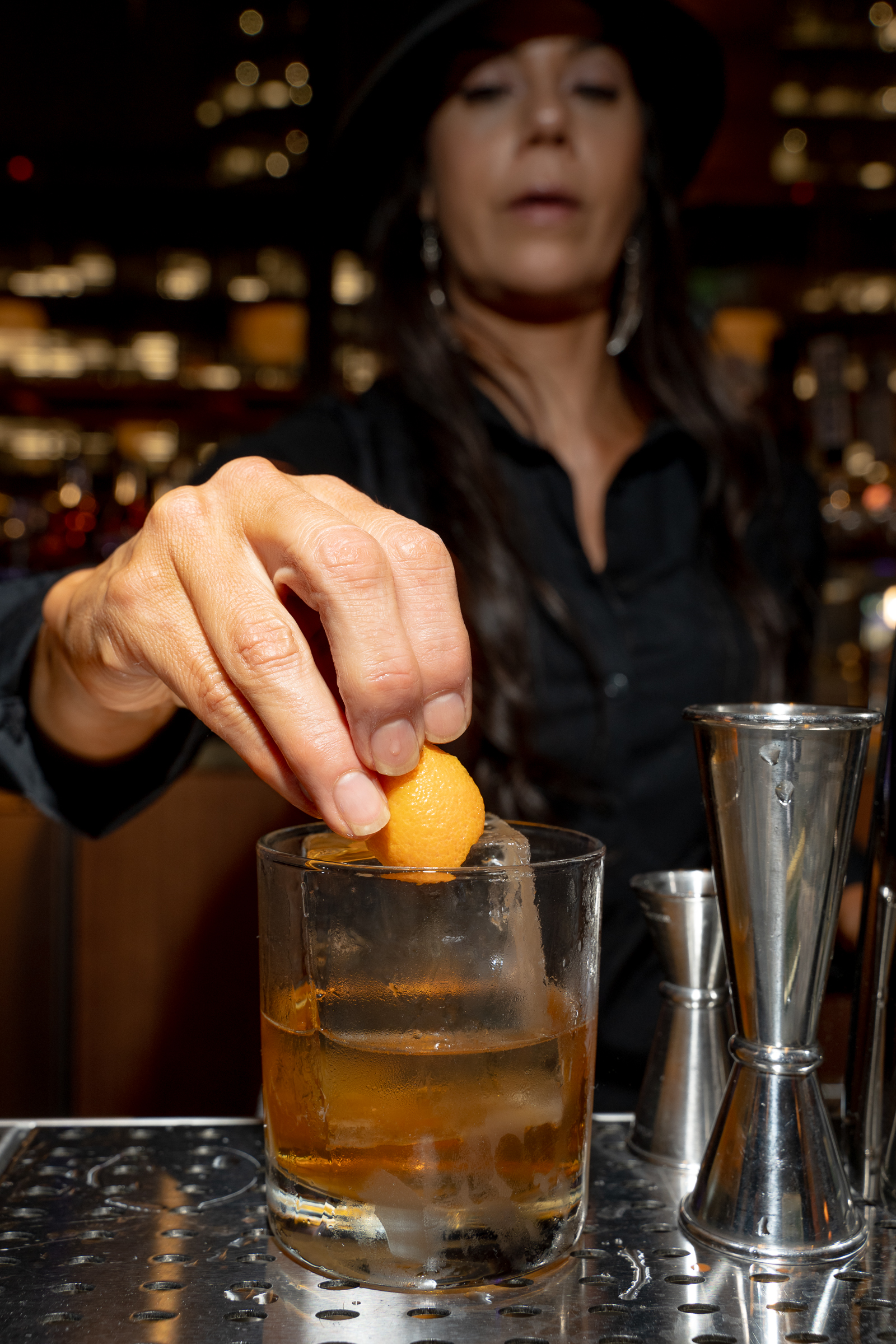 A person in a black shirt is squeezing a piece of orange peel over a glass with a brown drink and ice, next to a metal jigger on a bar counter.