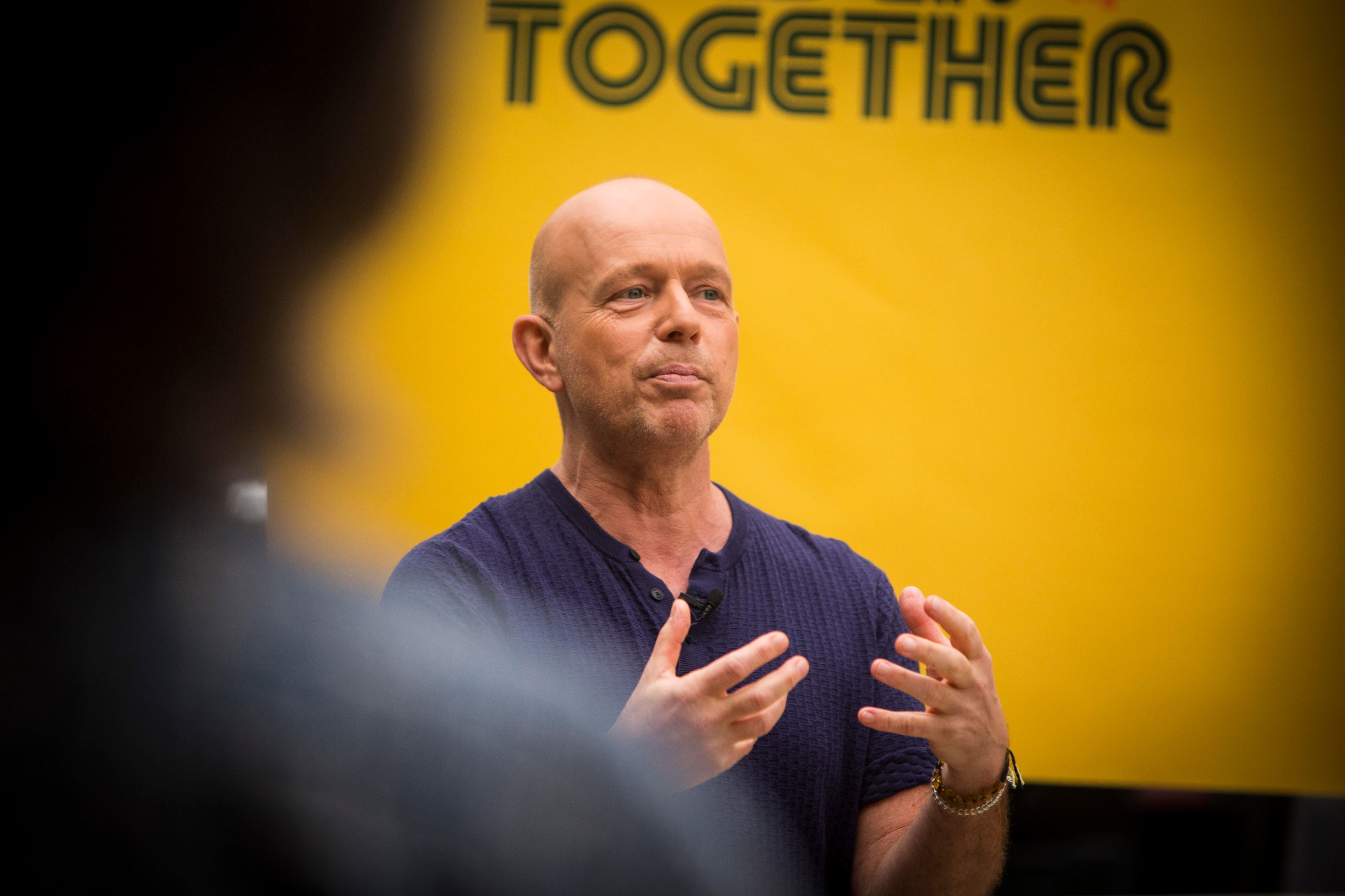 A bald man in a dark shirt gestures with his hands while speaking in front of a yellow background with partially visible text reading &quot;TOGETHER.&quot;