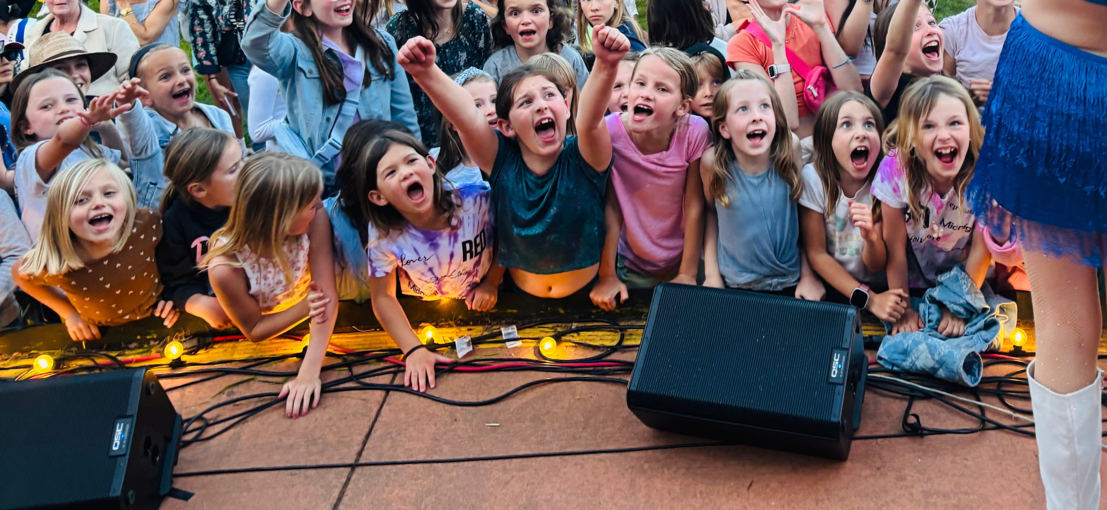 A group of excited children is cheering energetically at a concert. They're leaning over a barrier with expressions of joy and excitement.